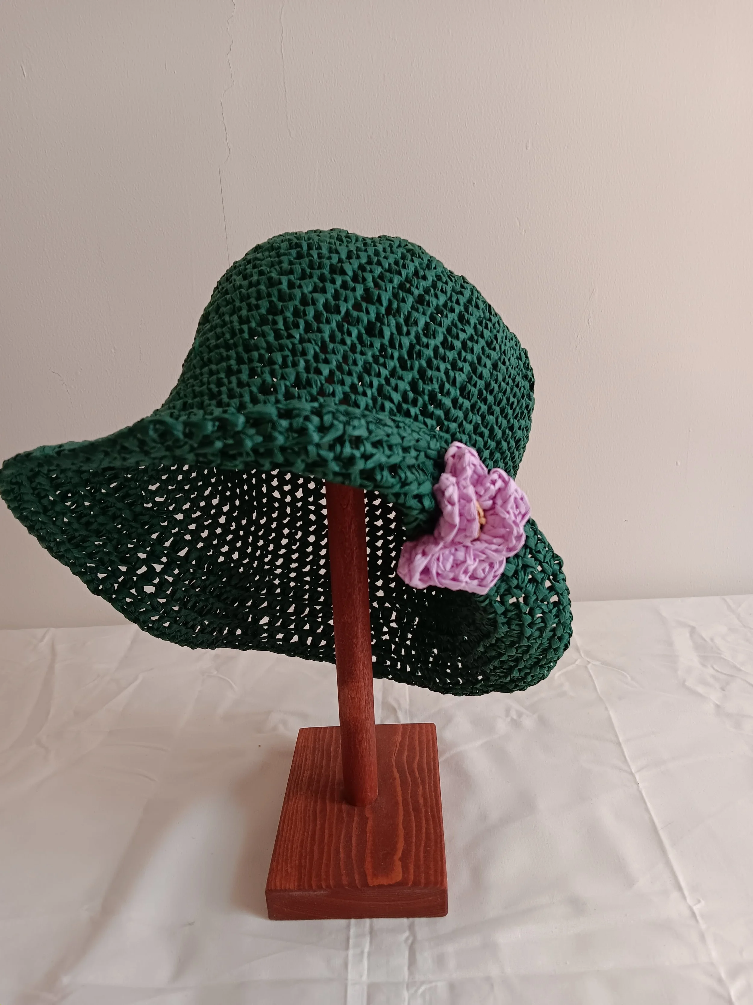 A decorative green straw sun hat with a pink fabric flower, mounted on a wooden stand against a plain white background.