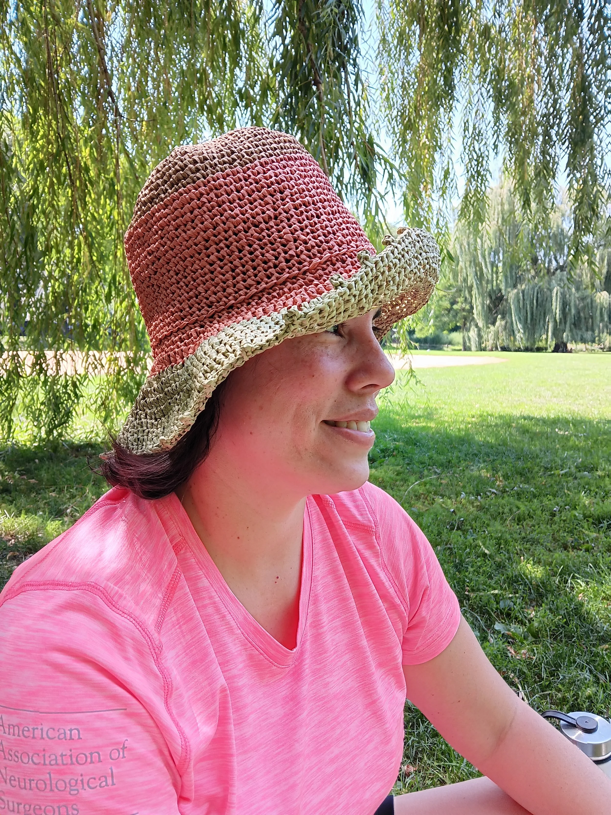 A woman with dark hair wearing a pink shirt and a wide-brimmed woven straw hat, sitting outdoors in a park with green grass and hanging tree branches in the background.