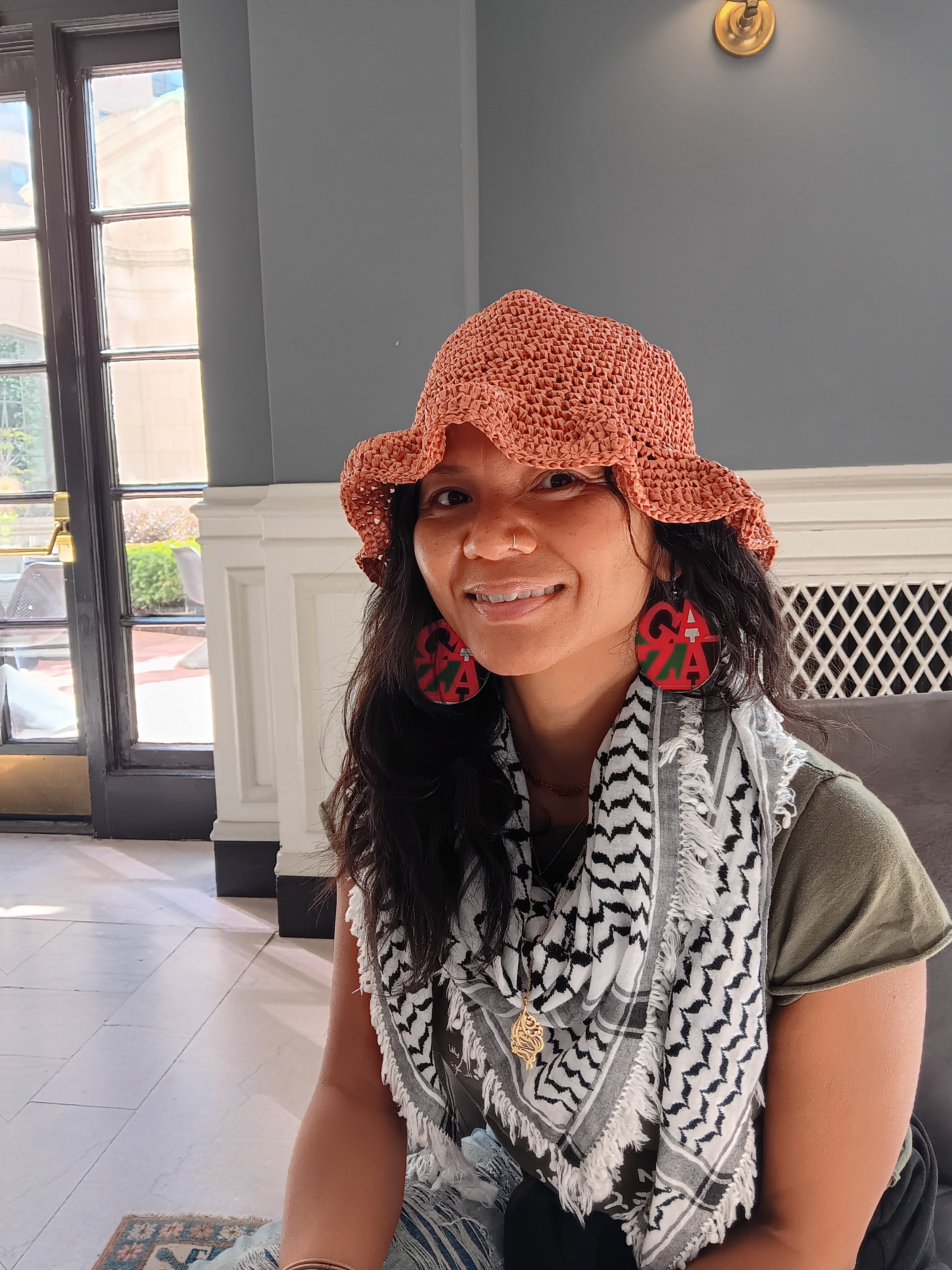 Woman wearing a large orange woven hat, red and green earrings, a gold necklace, and a black and white patterned scarf, sitting indoors near a window with natural light.