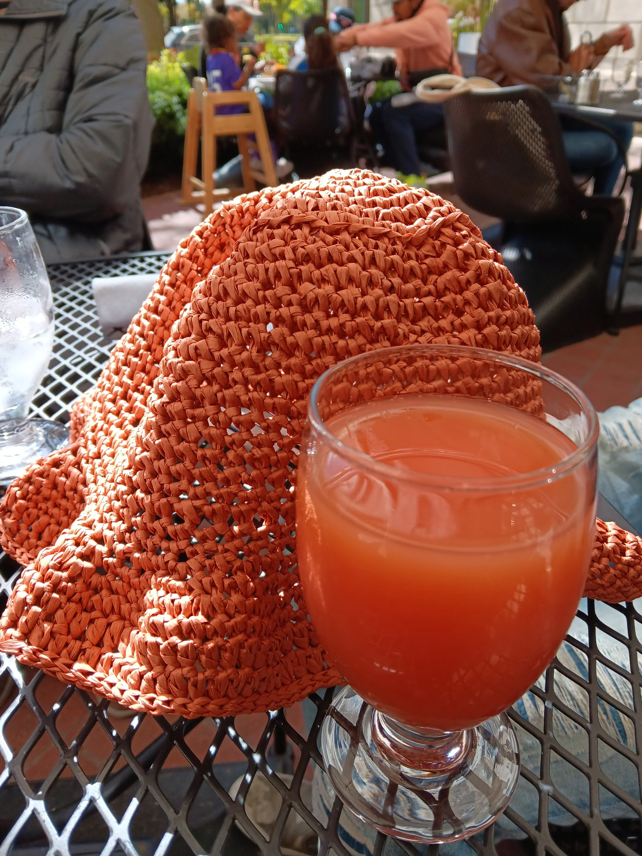 A woven straw hat, a glass of pinkish-orange drink, and a glass of water on a metal outdoor table, with people sitting and eating in the background at an outdoor cafe.