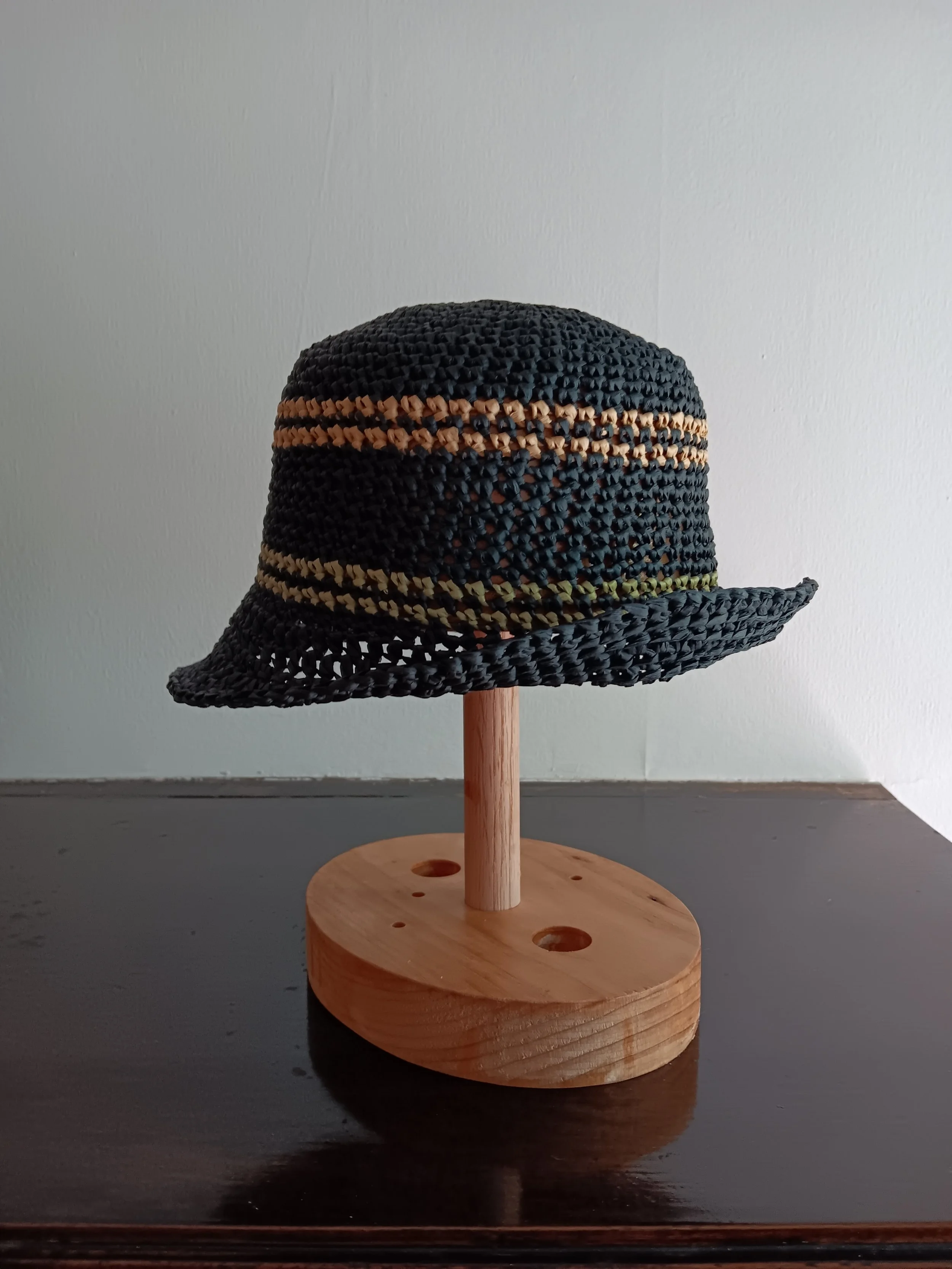 A woven straw hat with a wide brim, decorated with horizontal stripes in shades of beige and black, displayed on a wooden stand with a round base.