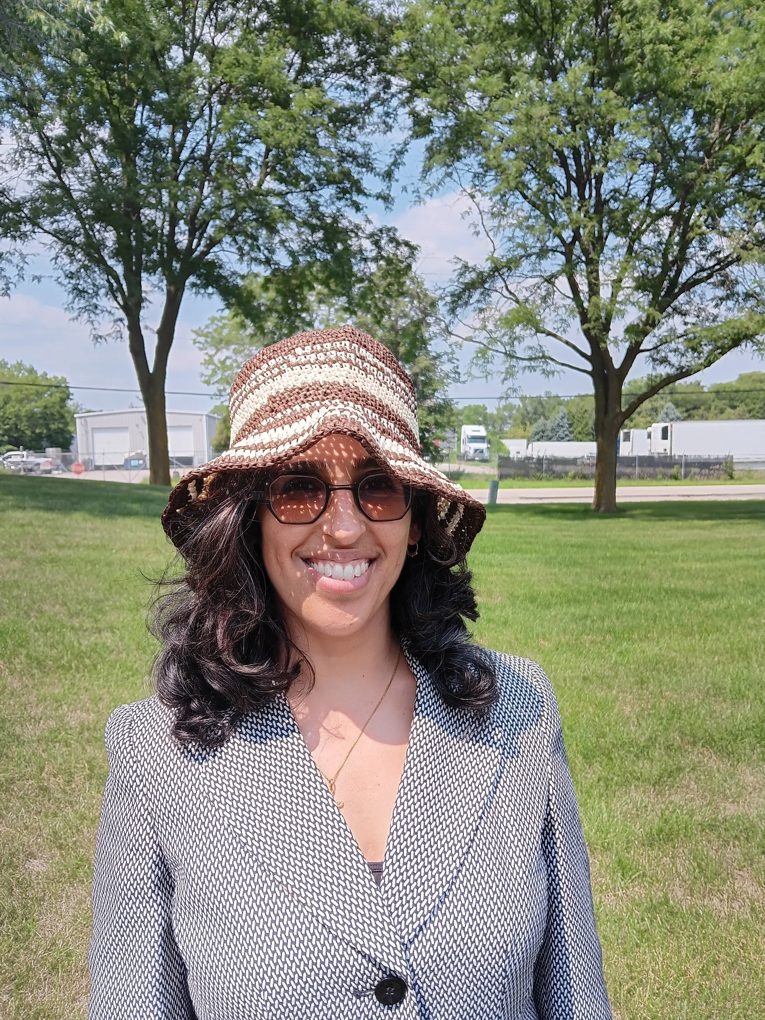 A woman smiling outdoors wearing a wide-brimmed hat, glasses, and a patterned blazer, with trees and a grassy area in the background.