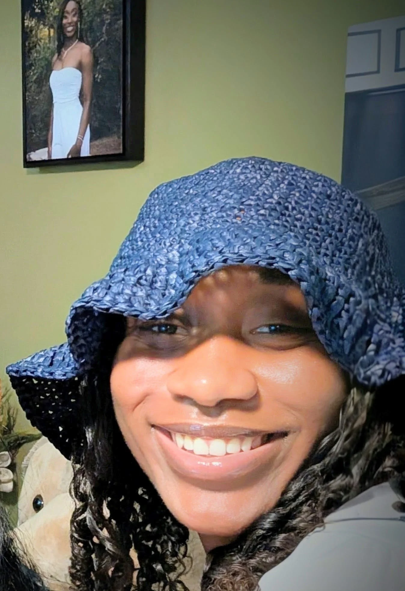 A person with dark curly hair smiling, wearing a large blue and white patterned cap, a beige jacket, and standing in front of a yellow wall with a framed photograph of a woman in a white dress.