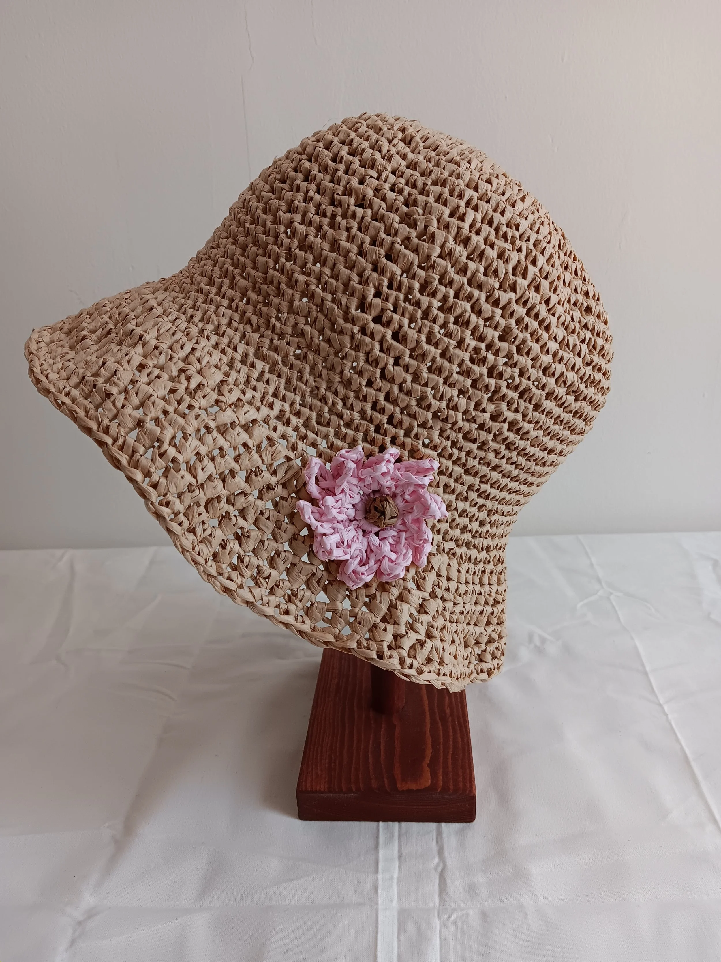 Woven straw hat with pink fabric flower and decorative brown flower, mounted on a wooden stand, with a plain wall background.