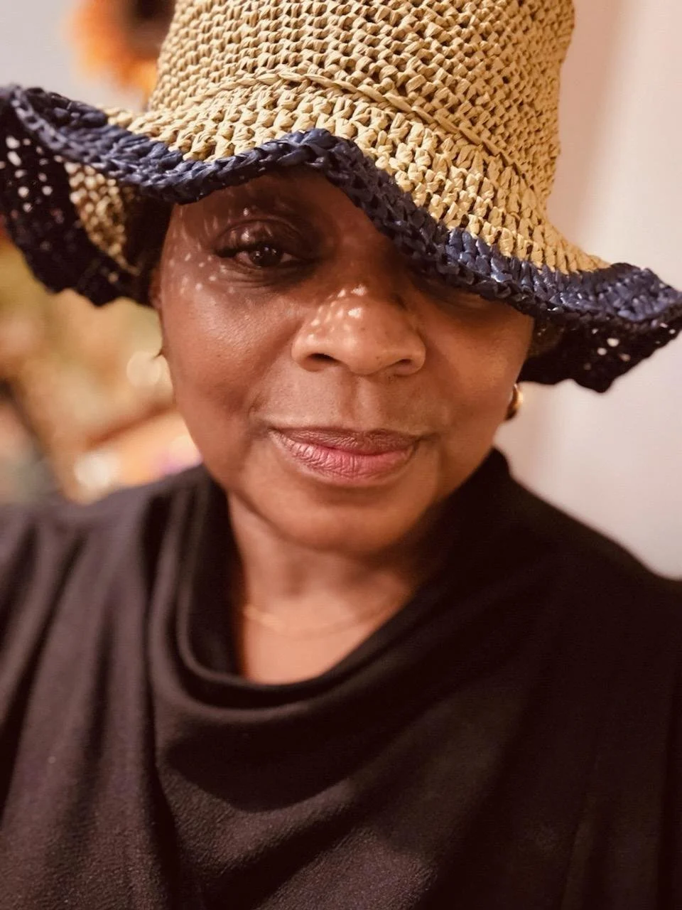 A woman wearing a large sun hat with a woven straw top and black woven brim, slightly tilting her head, wearing a black top, and smiling gently.