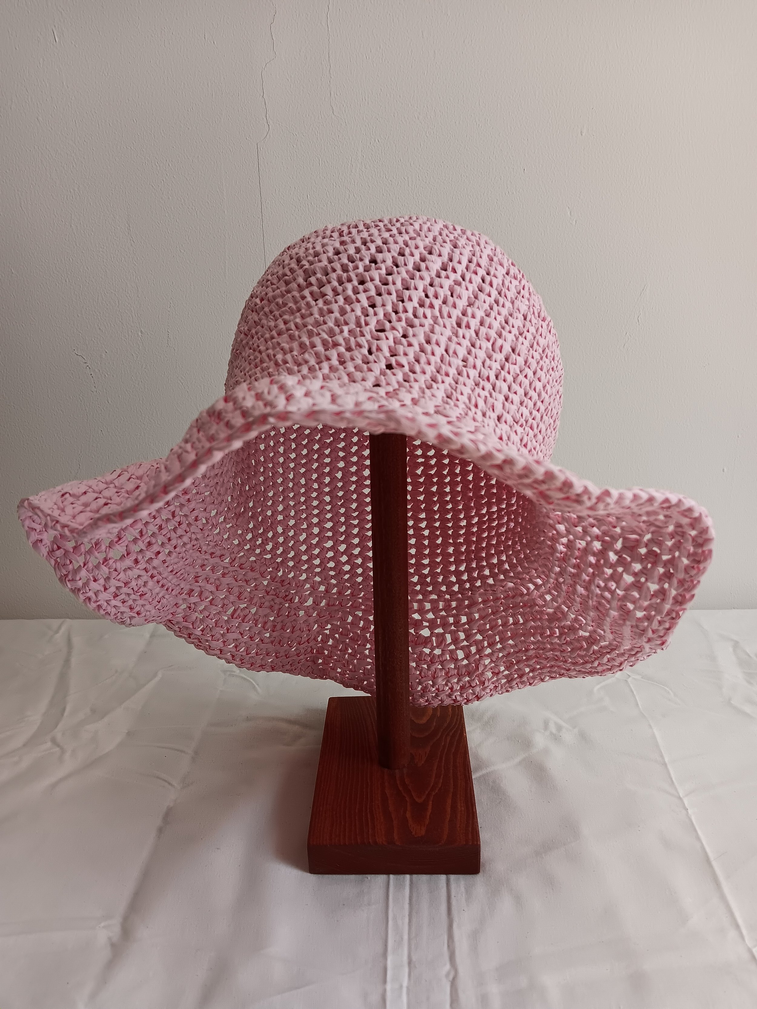 A pink woven sun hat on a wooden stand displayed on a white tablecloth, with a plain white wall in the background.