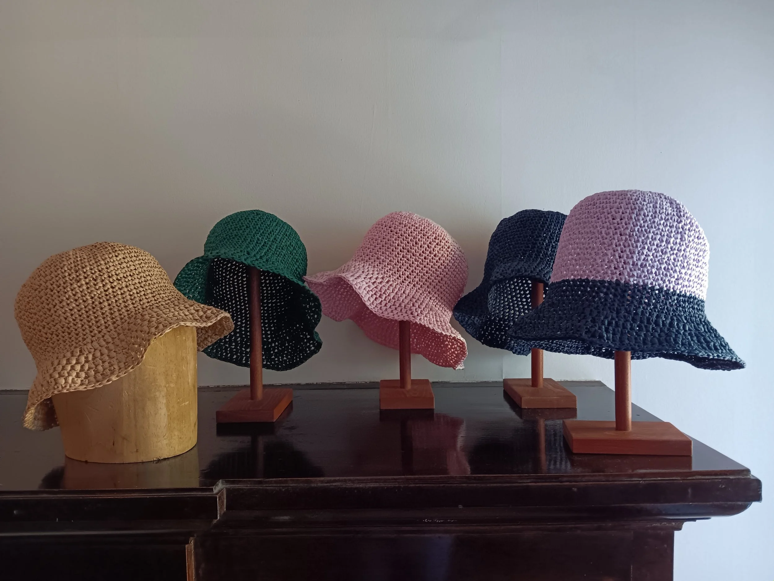 Display of colorful woven straw sun hats on wooden stands and a yellow wire hat on a wooden bust against a plain wall.