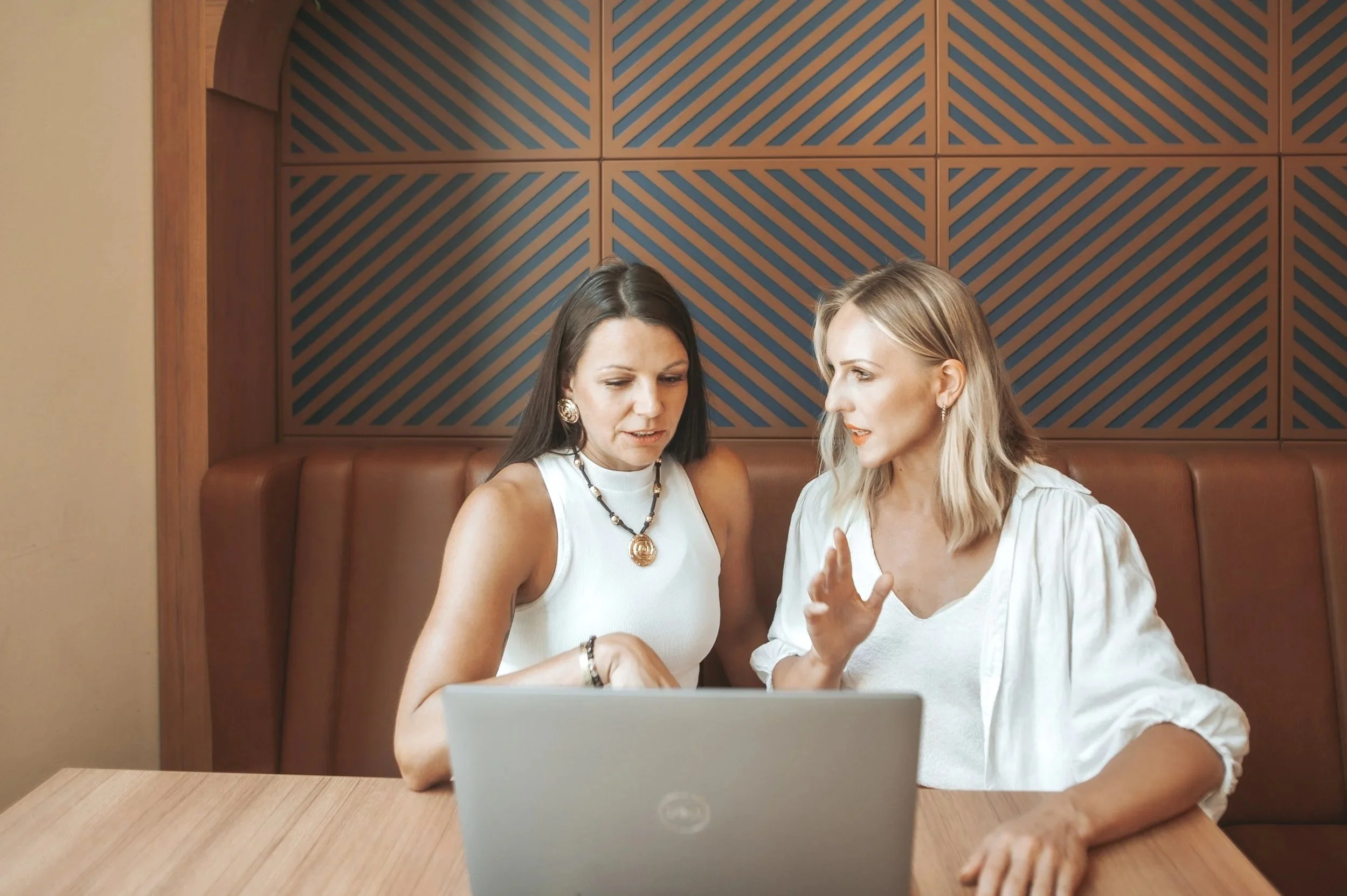 Zwei Frauen im Gespräch, sitzen an einem Tisch vor einem Laptop, hinter einer Wand mit Holzdesign.