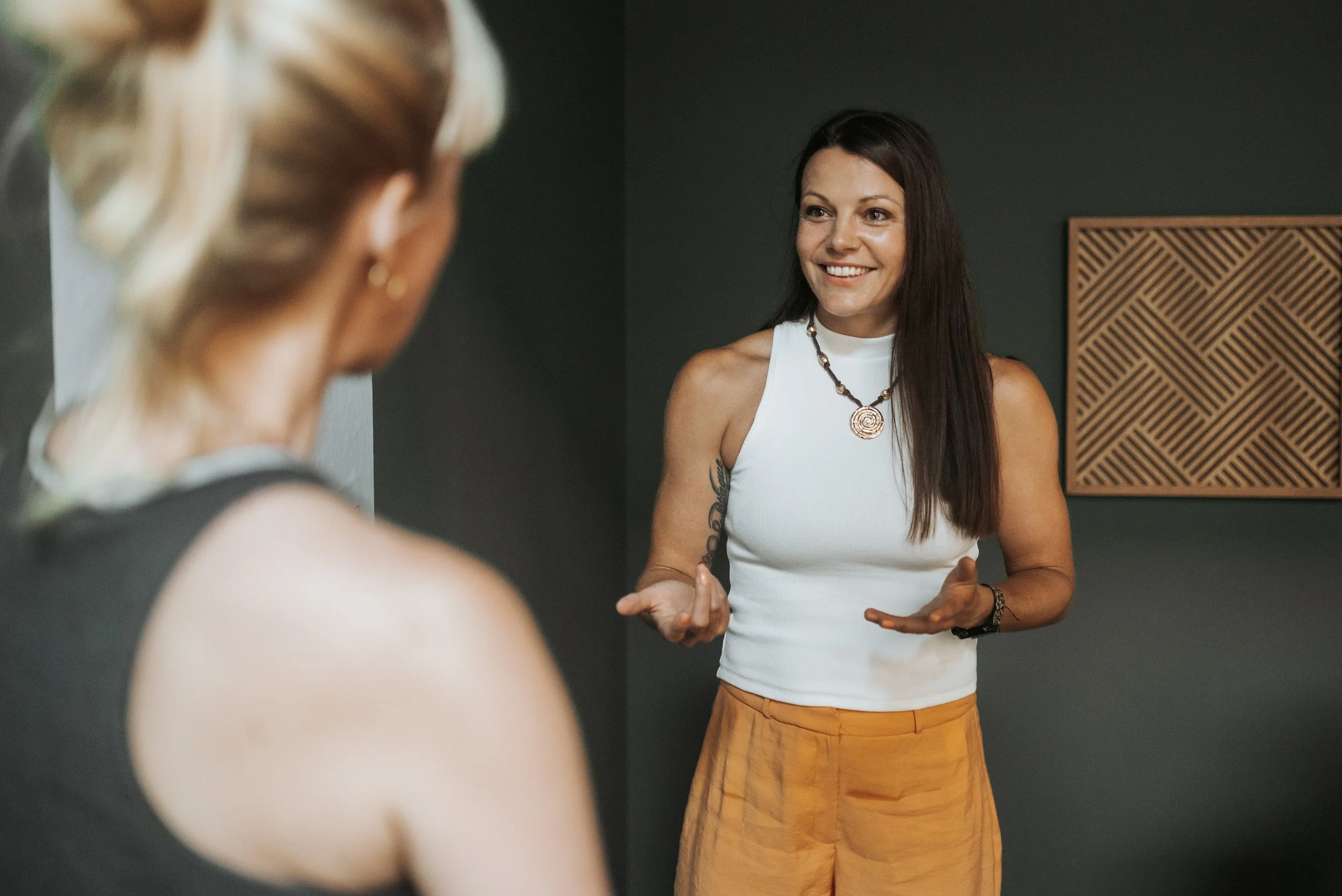 Zwei Frauen im Gespräch, eine im Vordergrund unscharf, die andere lächelnd, in einem Raum mit dunklen Wänden und Holzdekoration.