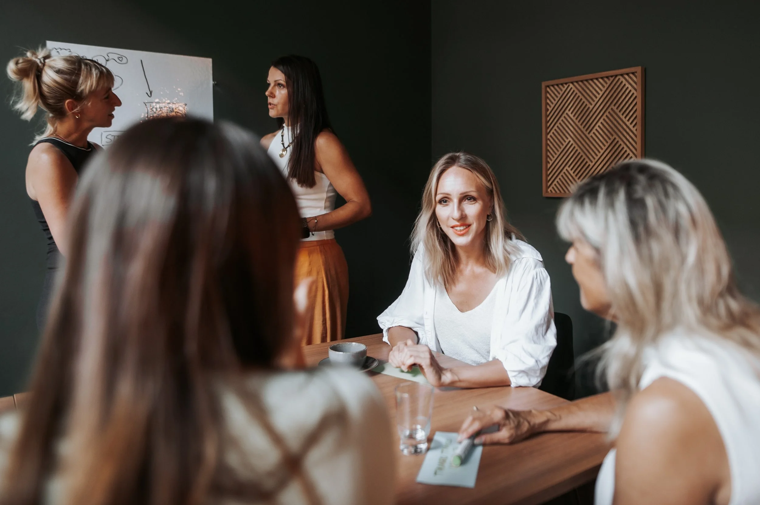 Eine Gruppe von Frauen sitzt an einem Holztisch in einem Raum mit dunklen Wänden. Zwei Frauen stehen vorne und sprechen sich an, während die anderen Frauen zuzuhören scheinen.