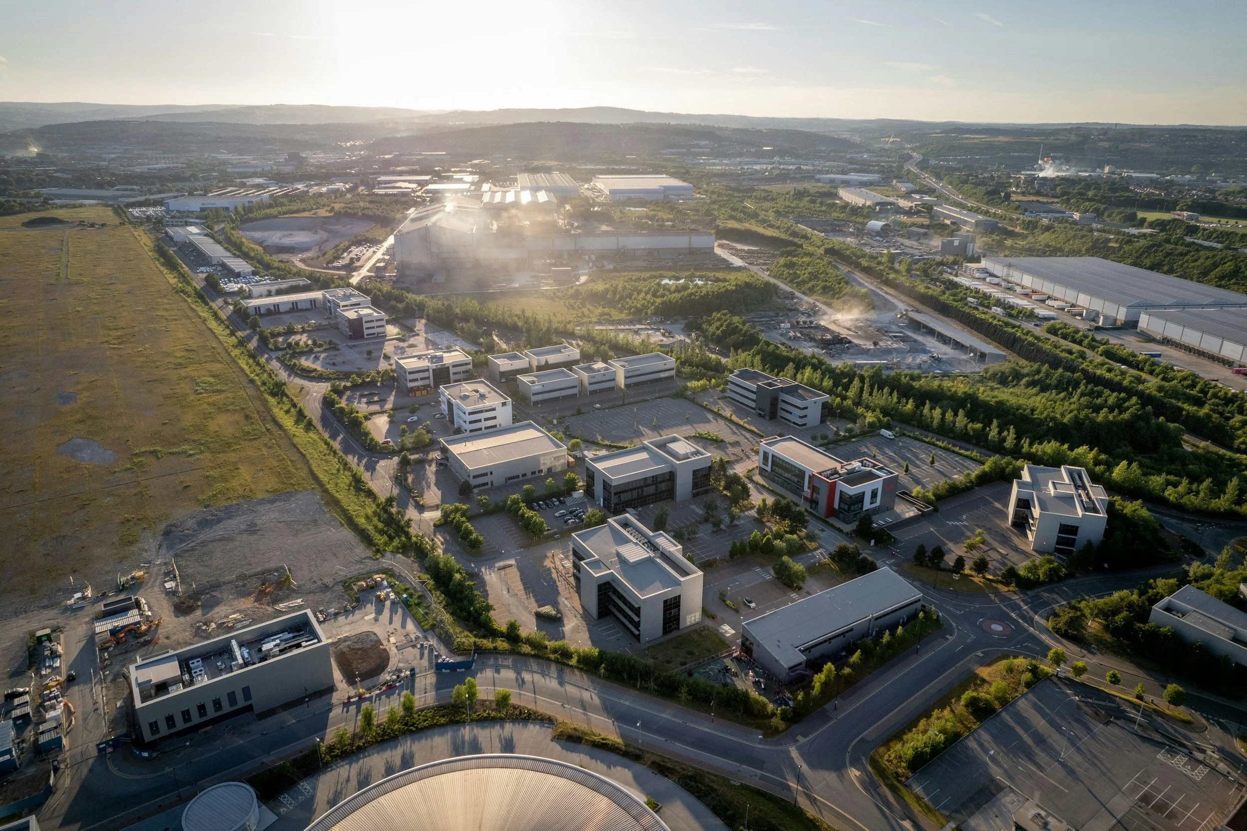 Aerial view of a cityscape with modern buildings, green spaces, parking lots, and some industrial areas, during late afternoon sunlight.
