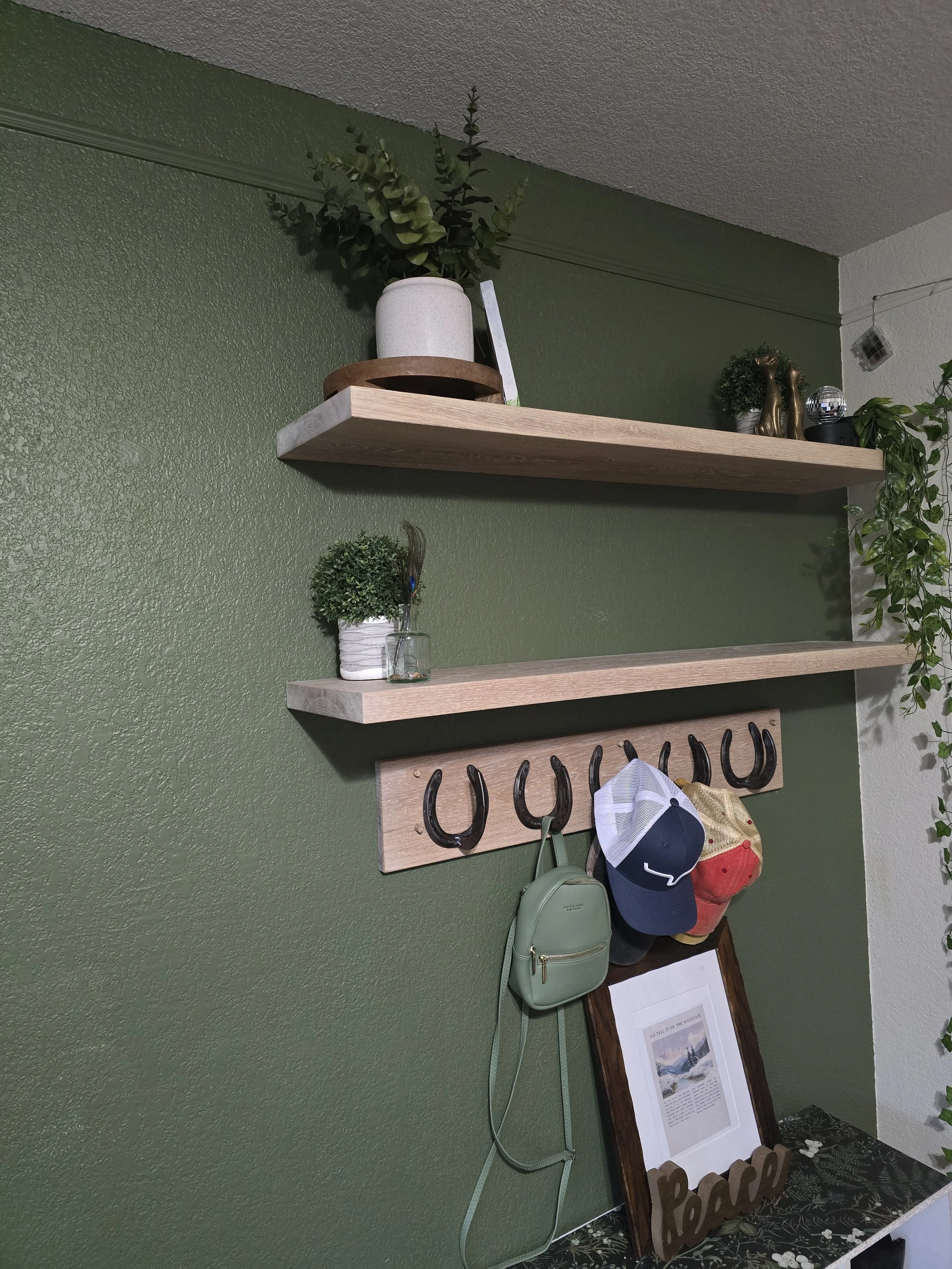 A hallway with a green painted wall and white ceiling, featuring two wooden shelves with decorative items and hooks holding hats and bags, along with an framed picture on a table below.