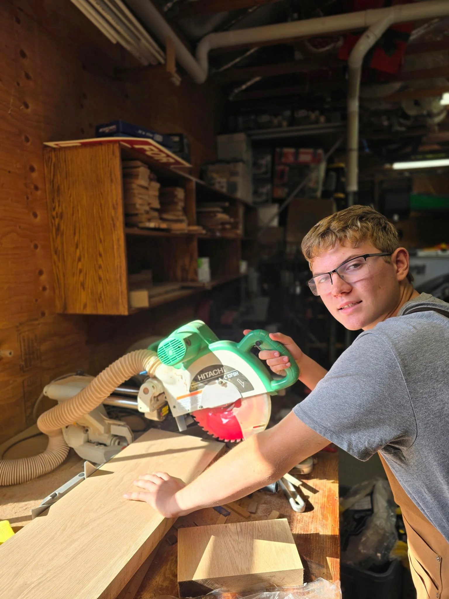 A young man with glasses cutting wood using a circular saw in a woodworking workshop.