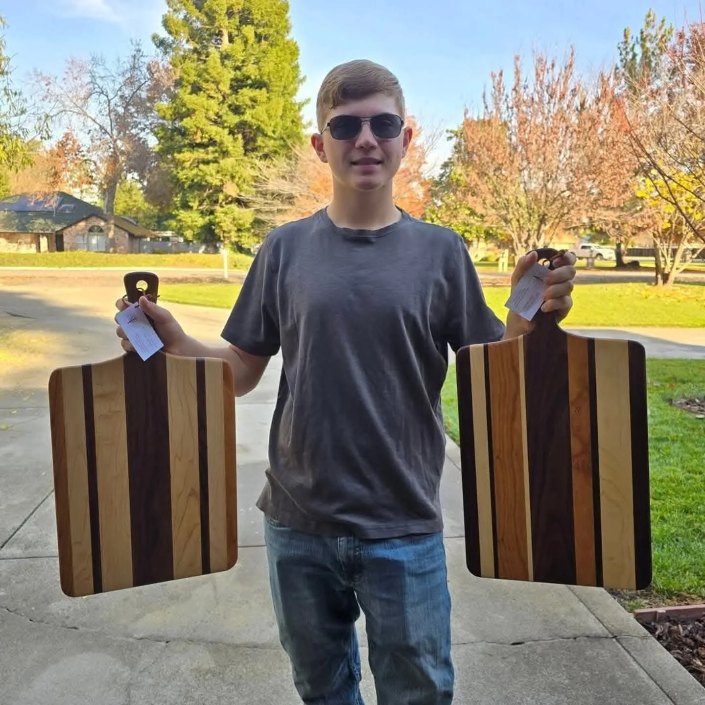 A young man wearing sunglasses and a gray t-shirt holding two wooden cutting boards outdoors on a sidewalk with trees and houses in the background.