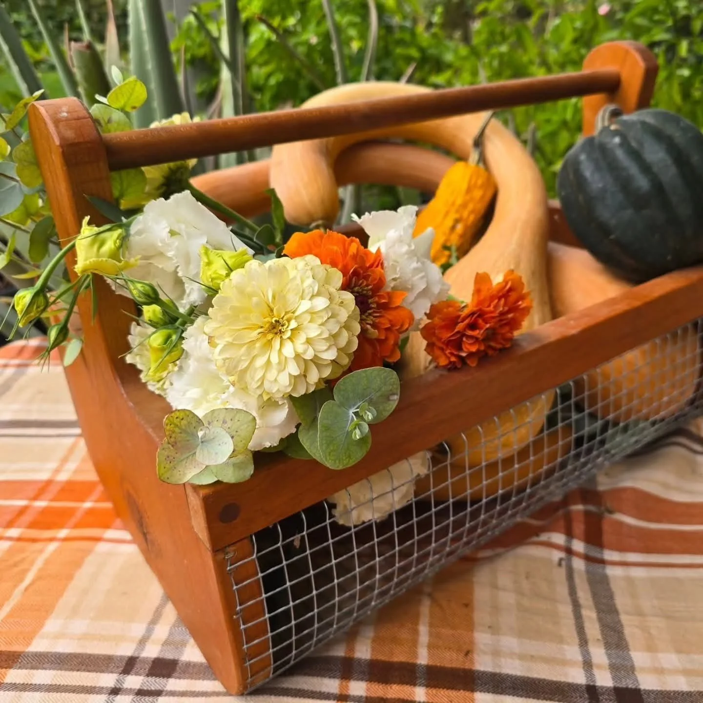 A wooden basket filled with colorful flowers, including white, orange, and yellow blossoms, alongside green leaves, on a checkered tablecloth.