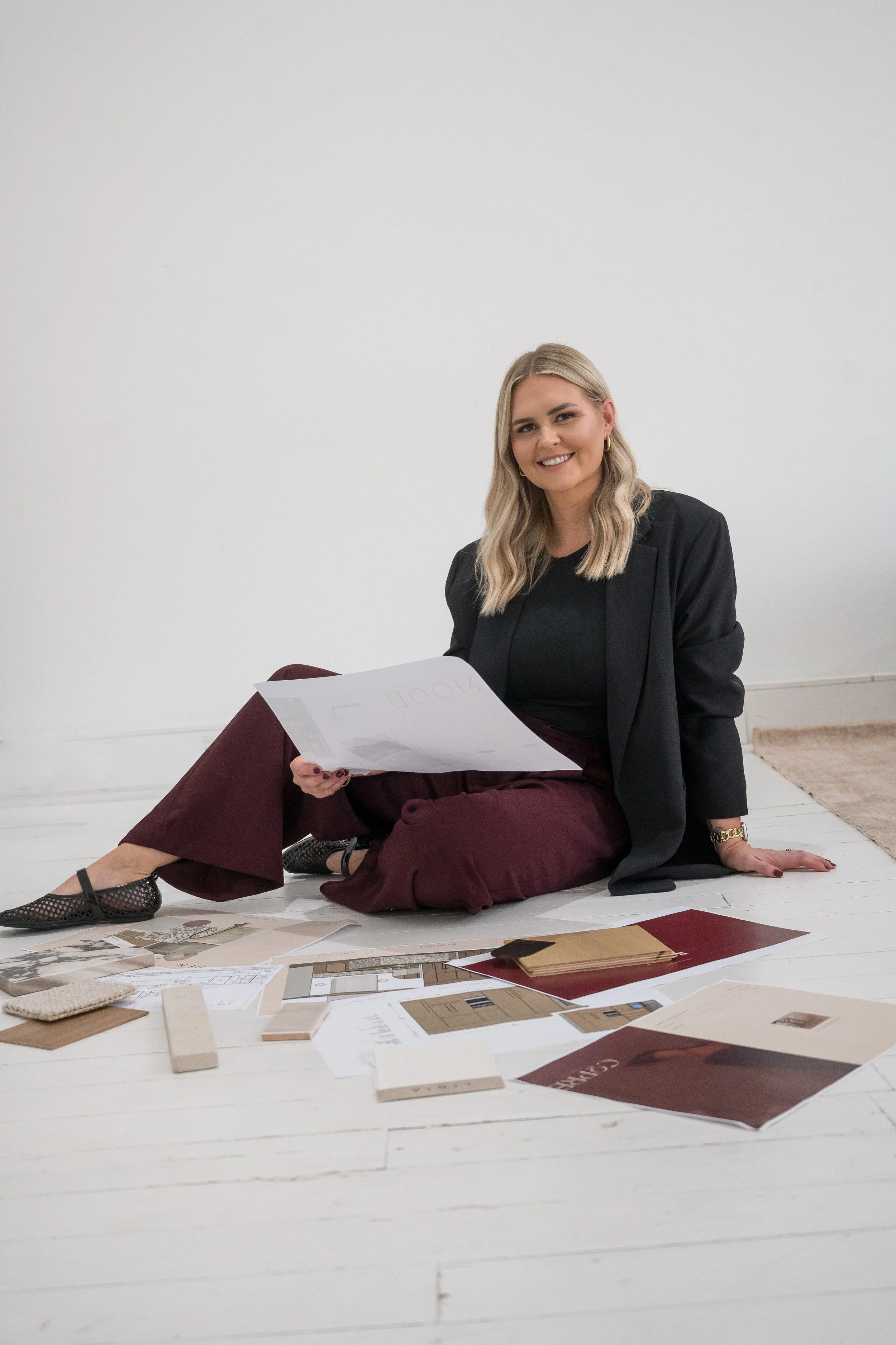 A woman with blonde hair sitting on the floor, smiling, surrounded by design and interior materials including fabric samples, color swatches, and printed flooring or wall design plans.