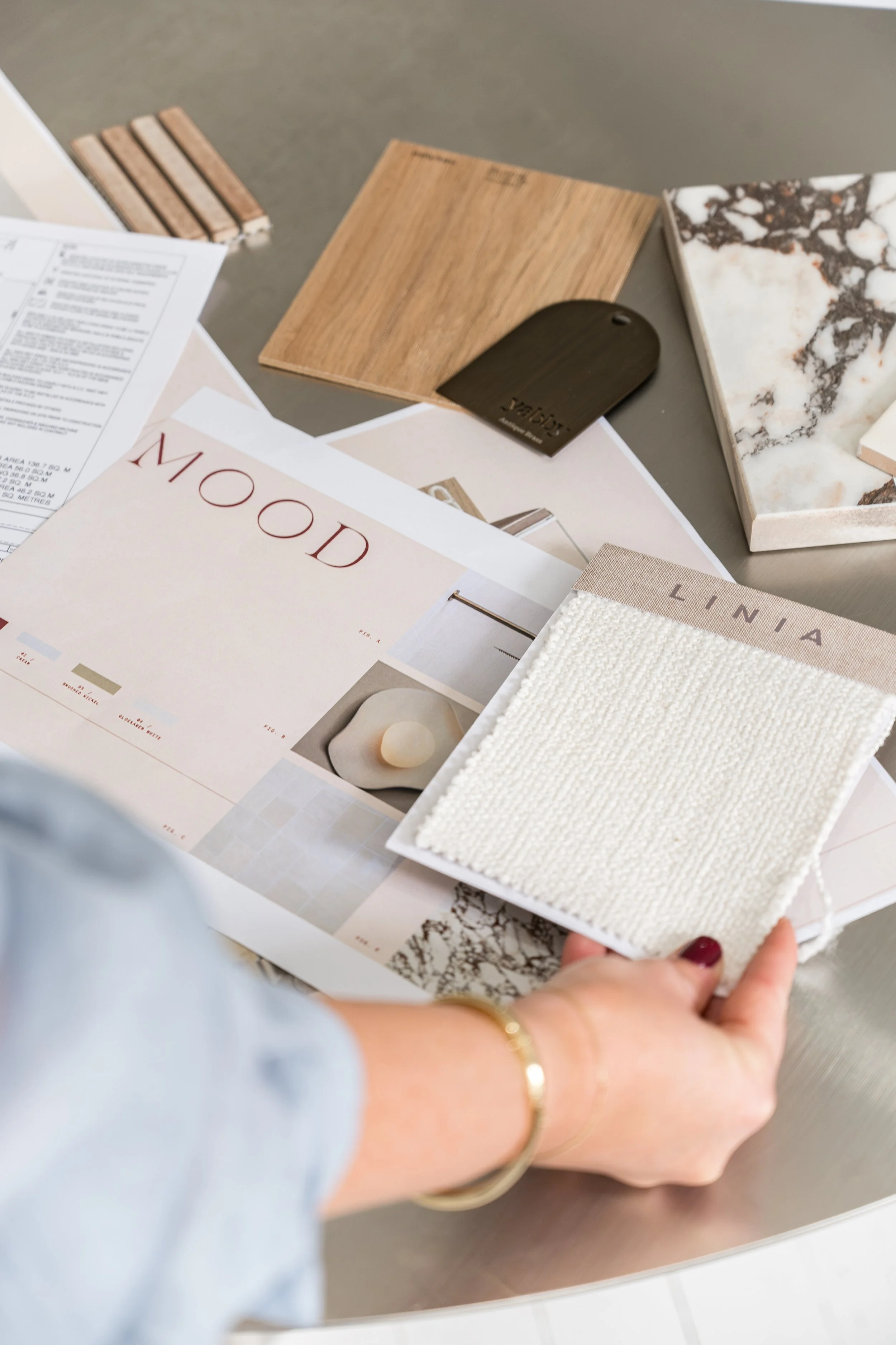 A person with red nail polish holding a small fabric swatch with a textured white fabric sample. In the background, there are samples of wood, marble, and a printed design sheet on a table.