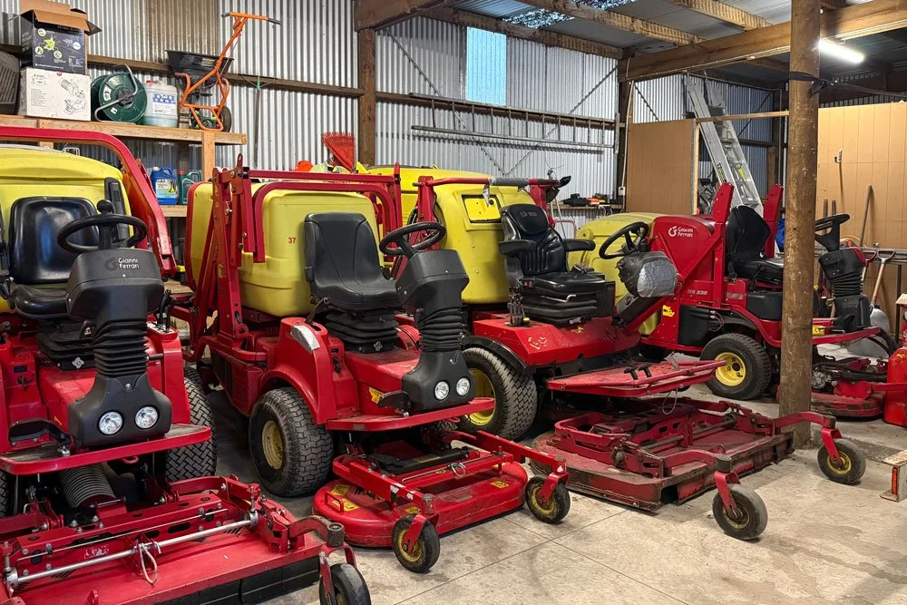 Multiple red and yellow ride-on lawn mowers are parked inside a barn with corrugated metal walls and wooden supporting beams.