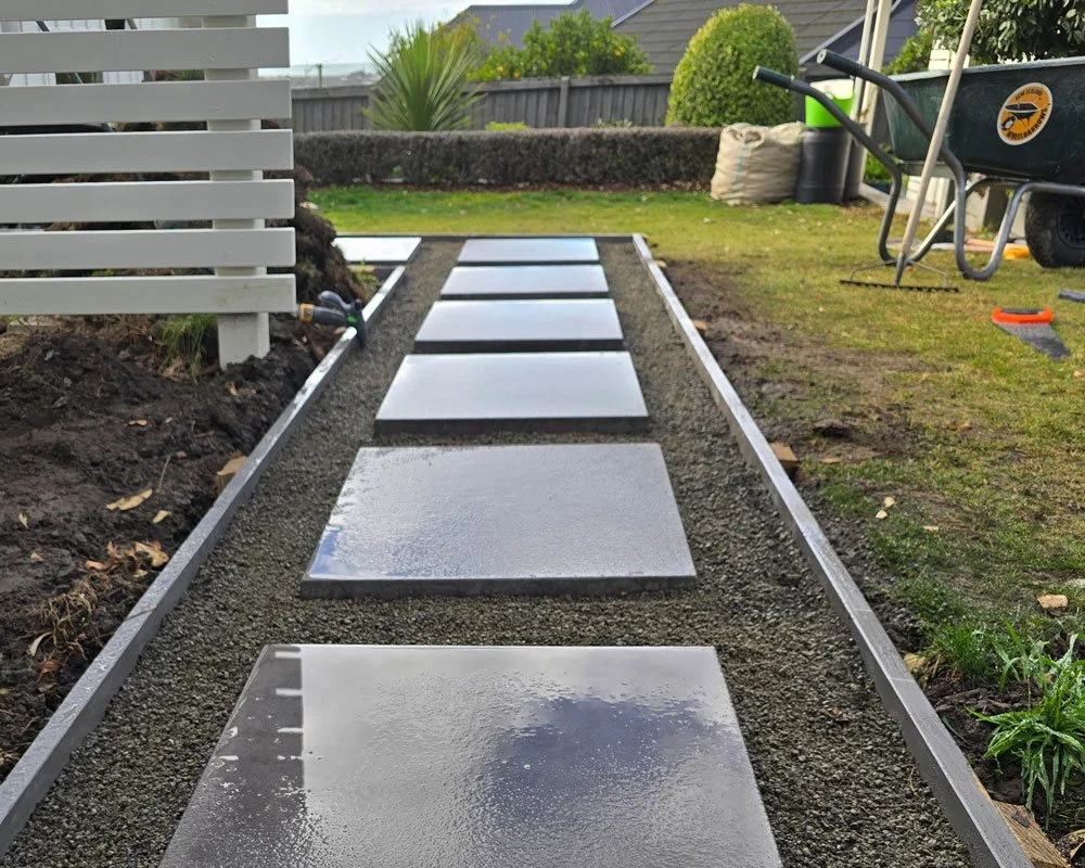 Wet concrete pathway with square stepping stones in a backyard, with gardening tools and wheelbarrow nearby.