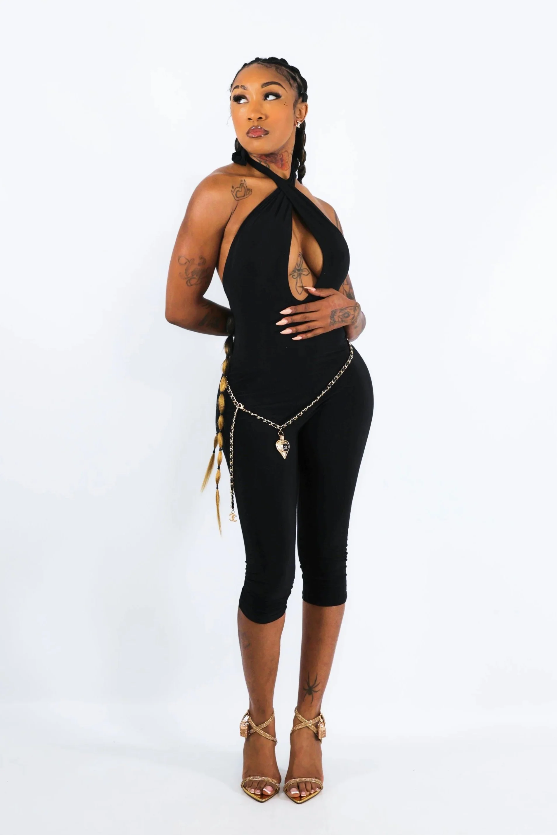 A woman with braided hair and tattoos is wearing a black halter top, black pants with chains, and gold high heels against a plain white background.