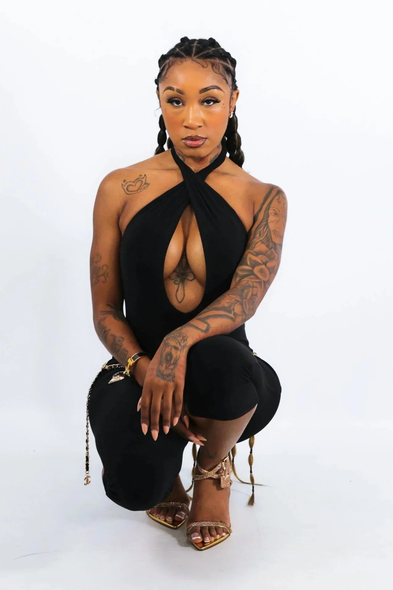 A woman with tattoos on her arms and chest wearing a black halter top with a deep neckline, black pants, and gold high-heeled sandals, sitting against a plain white background.