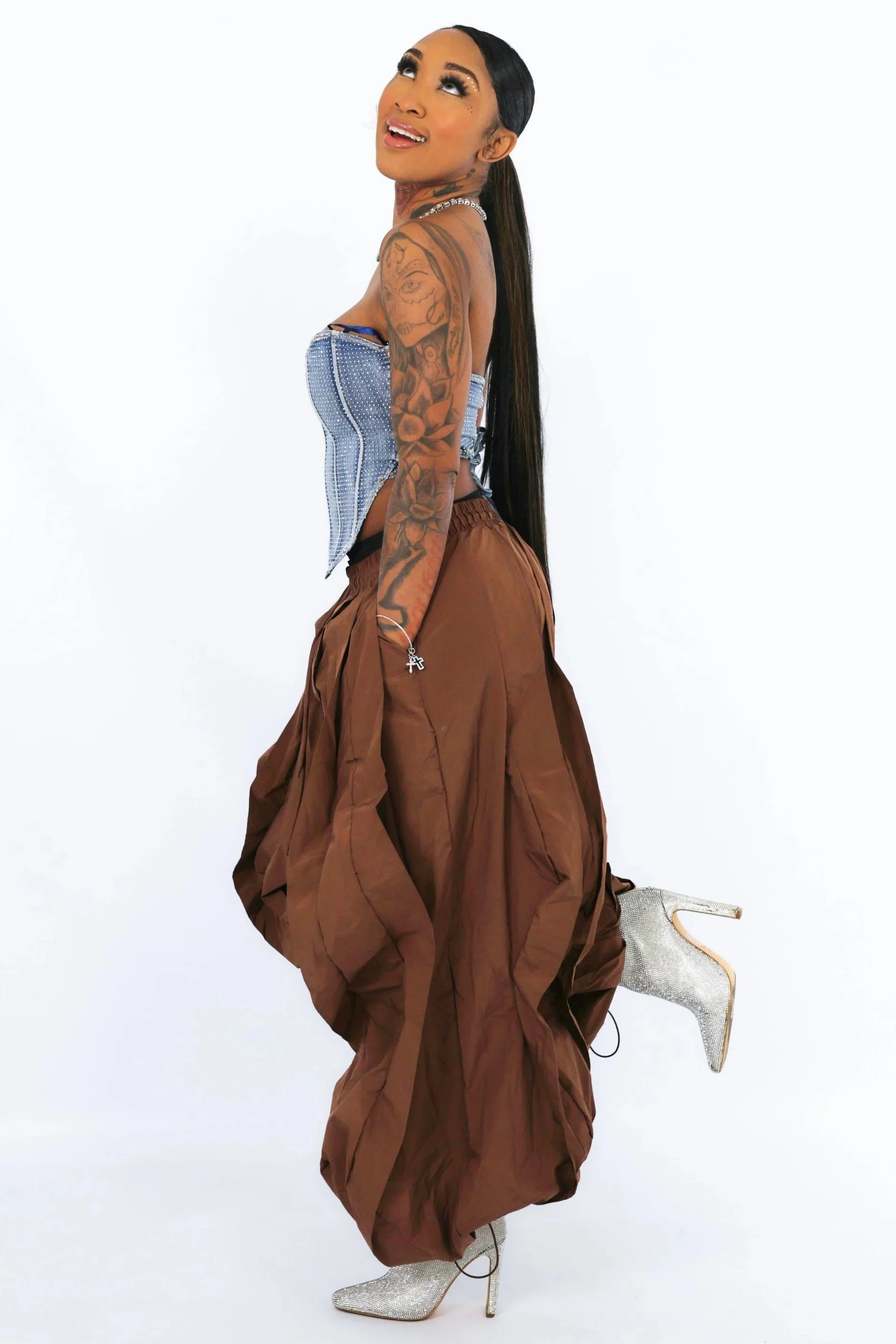 A woman with long dark hair, tattoos on her arms and face, wearing high heels, a beaded necklace, and brown puffed pants. She is posing against a plain white backdrop, looking upward with a bright smile.