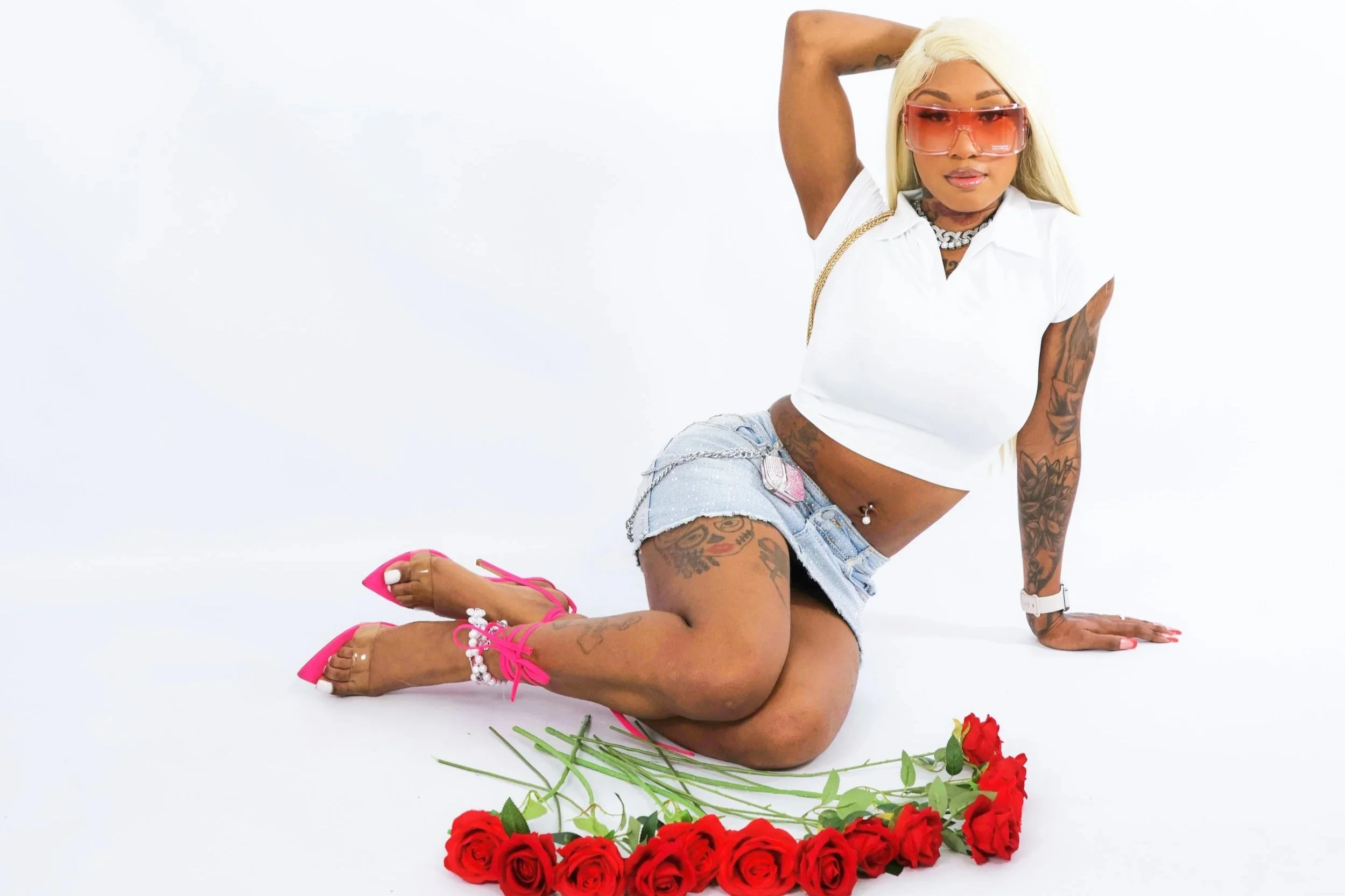 A woman with blonde hair and tattoos is on her hands and knees on a white surface, surrounded by red roses. She is wearing large pink sunglasses, a white cropped shirt, distressed denim shorts, and pink high-heeled shoes. The woman has one arm raised behind her head and is looking at the camera.