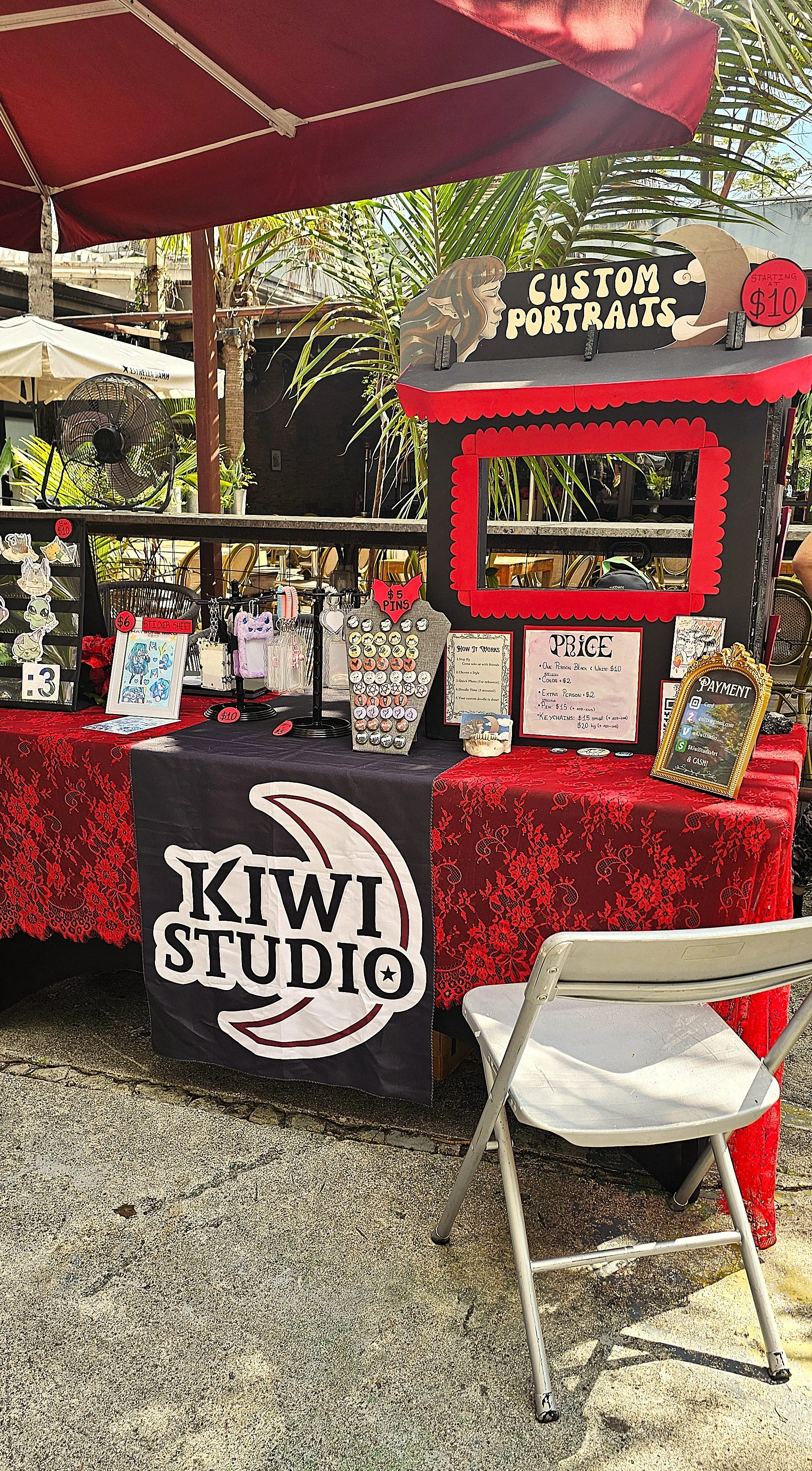 Outdoor booth selling custom portraits with a sign, price list, and various small items, featuring a red and black color scheme and a Kiwi Studio banner, set under red umbrellas and surrounded by greenery.
