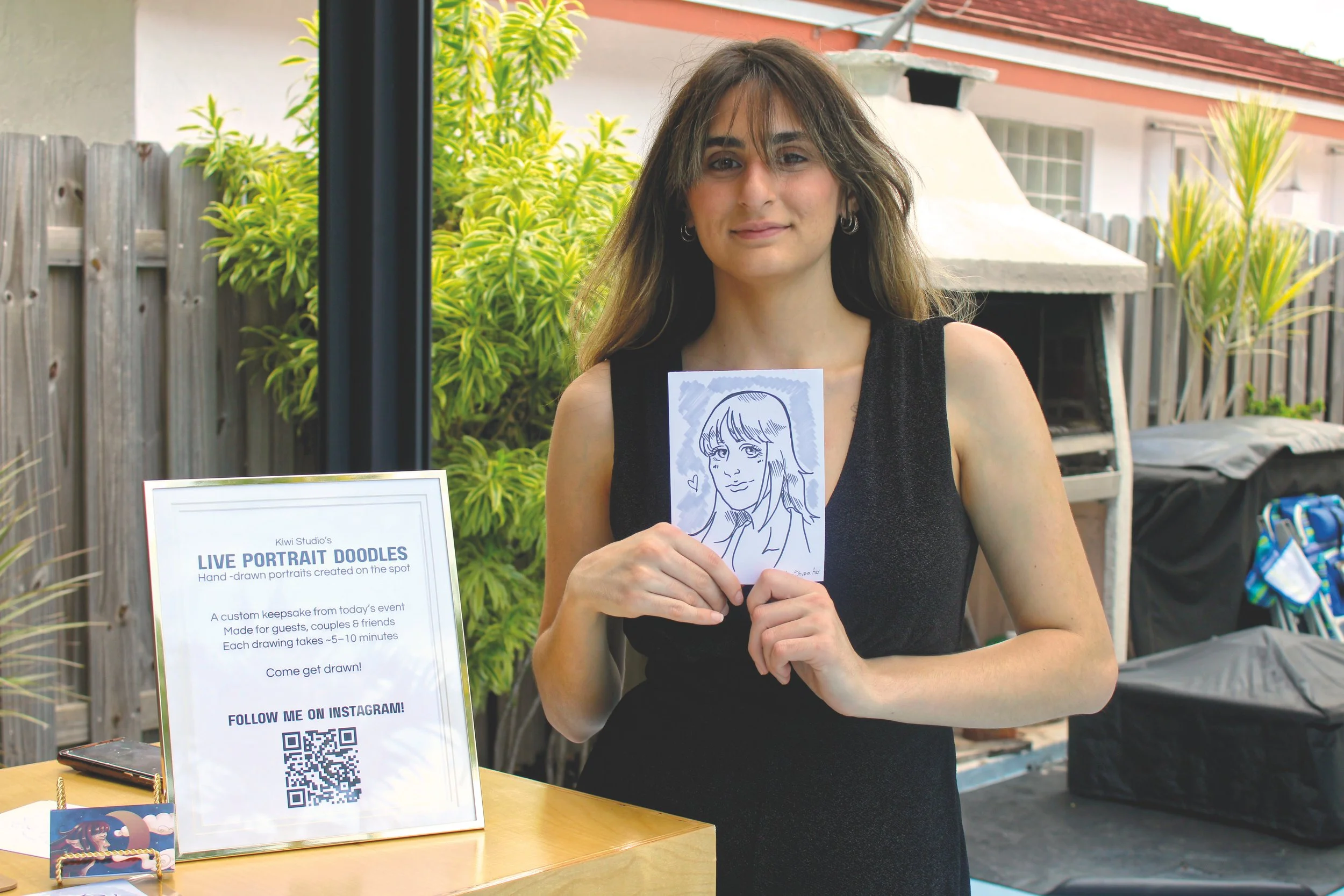 A woman with long brown hair and hoop earrings, wearing a black sleeveless top, holding a hand-drawn portrait of herself at an outdoor art event. There is a sign beside her promoting live portrait drawings with a QR code, and a table with art supplies.