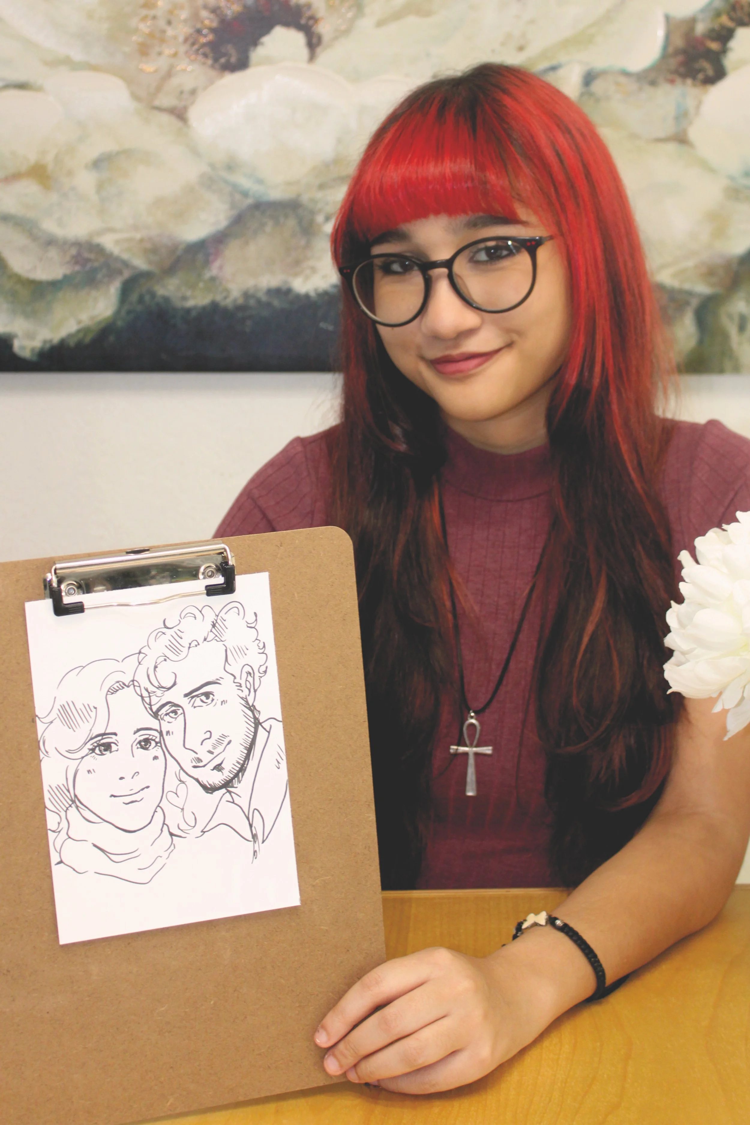 A woman with red hair and glasses sitting at a table, with a drawing of a woman and a man on a clipboard in front of her she made. She is smiling slightly and wearing a burgundy top, a black bracelet, and a necklace with an ankh pendant.