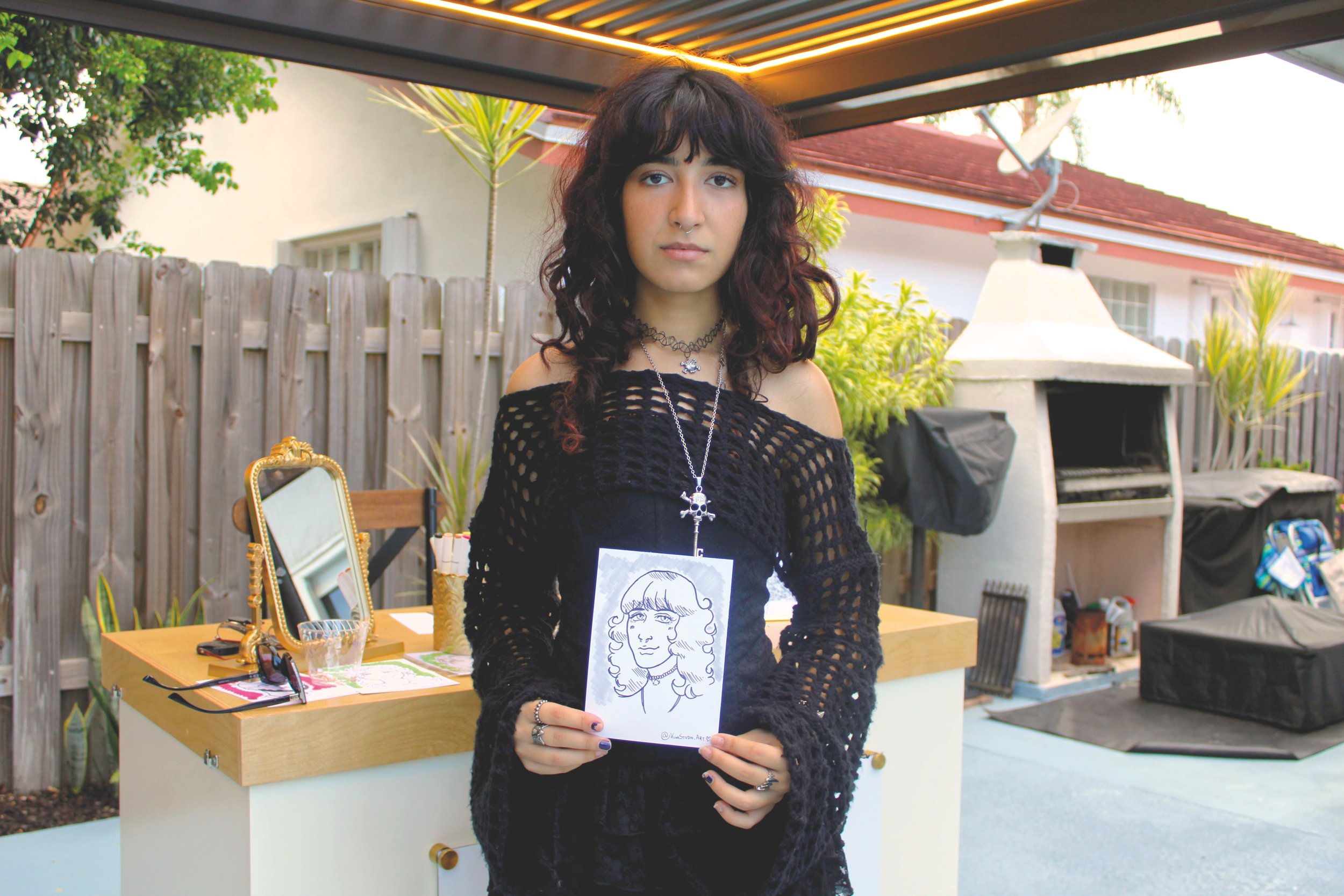 A young woman with dark curly hair holding a drawing of herself, standing outdoors behind a table with art supplies.