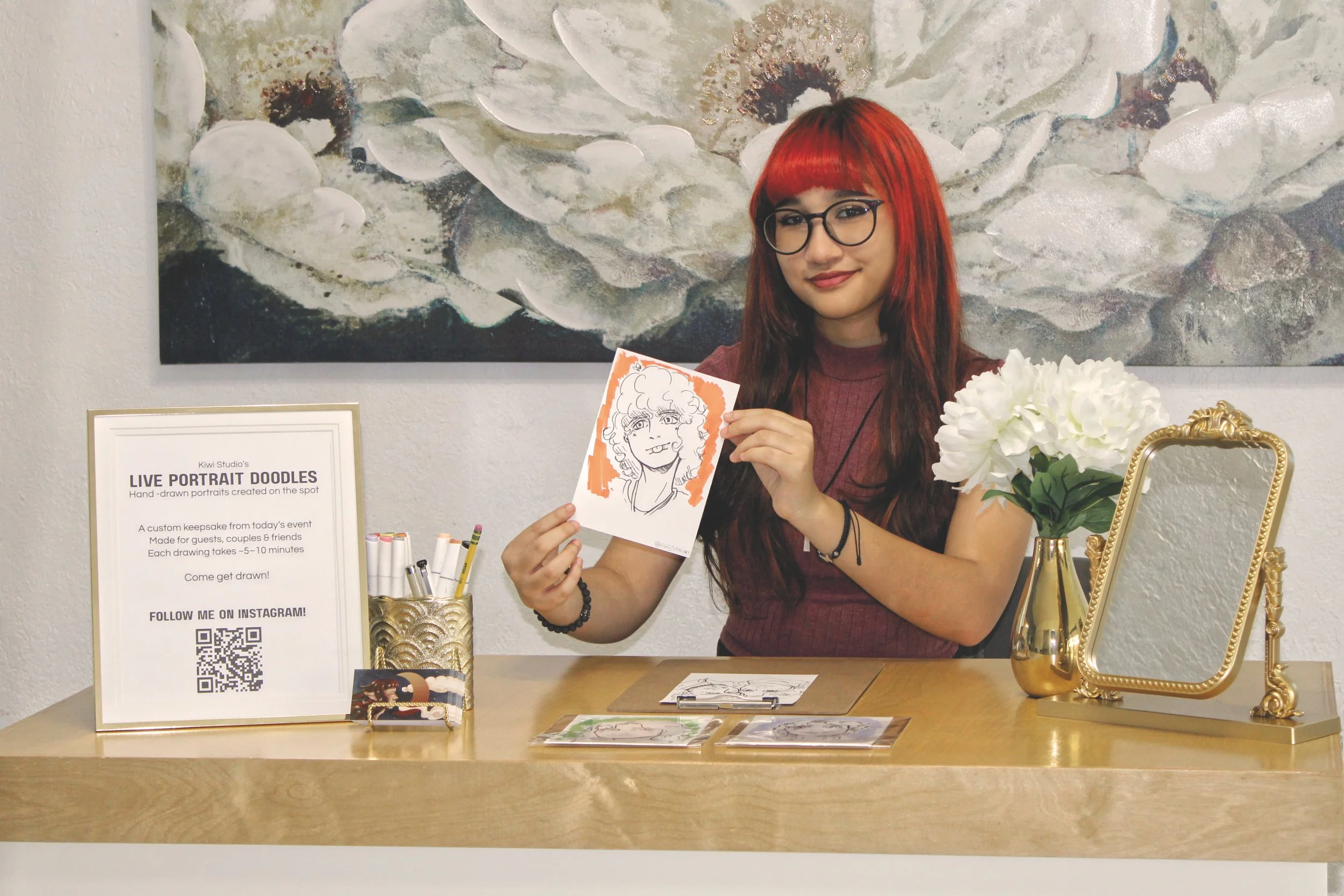 A woman with red hair and glasses holding a hand-drawn portrait of herself at an art event, with signs and art supplies on the table, and a large floral artwork on the wall behind her.