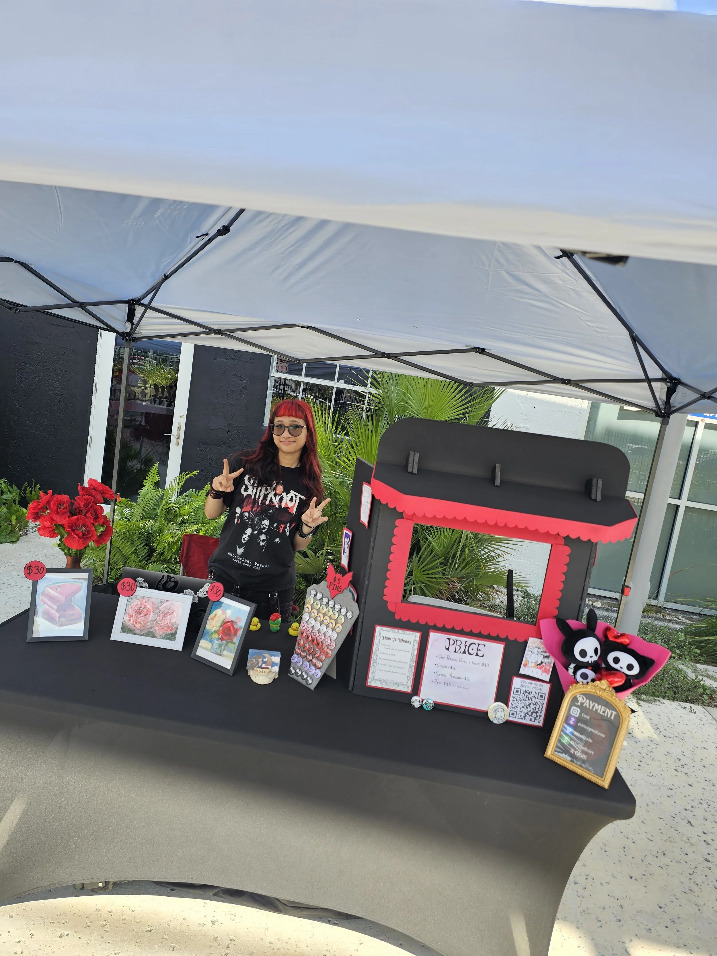 A woman with red hair and sunglasses standing behind a art booth at an outdoor market. The booth displays framed artwork, small figurines, and plush toys with skull characters. There are signs showing prices, and a canopy overhead. Behind her are plants and a building with large windows.