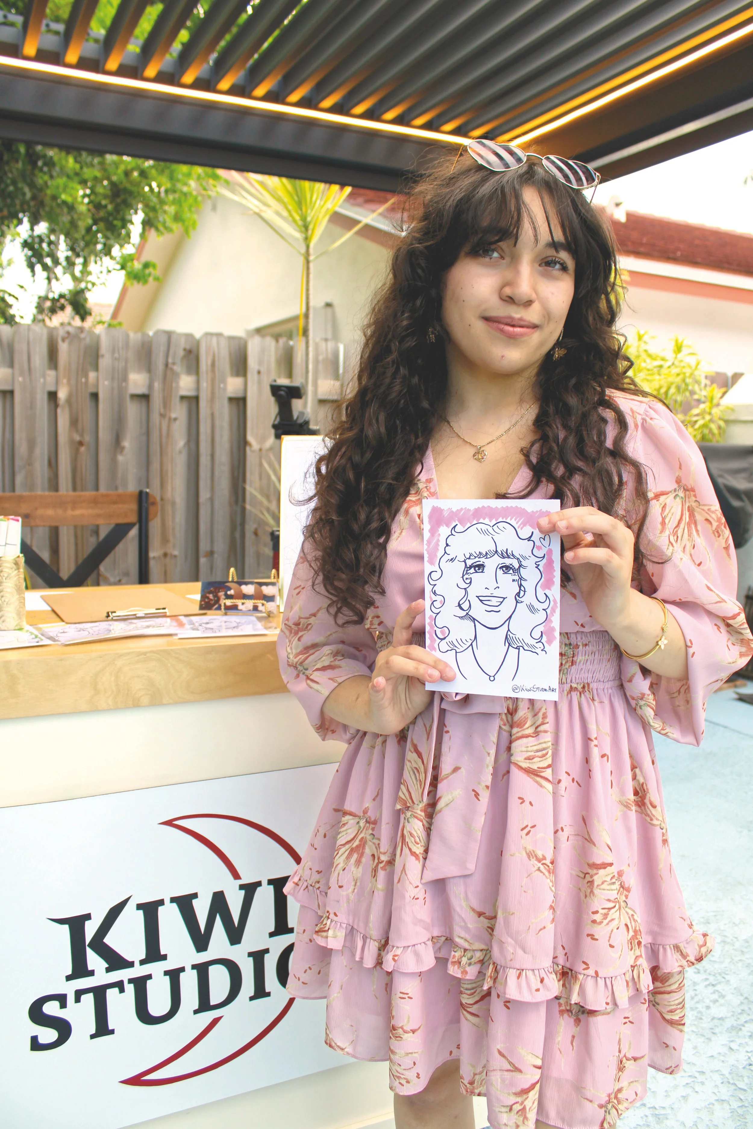 A woman with long curly dark hair and sunglasses on her head, smiling and holding a caricature drawing of herself, standing outdoors near a table with art supplies, in front of a sign that reads 'KIWI STUDIO.'; the booth of the artist that