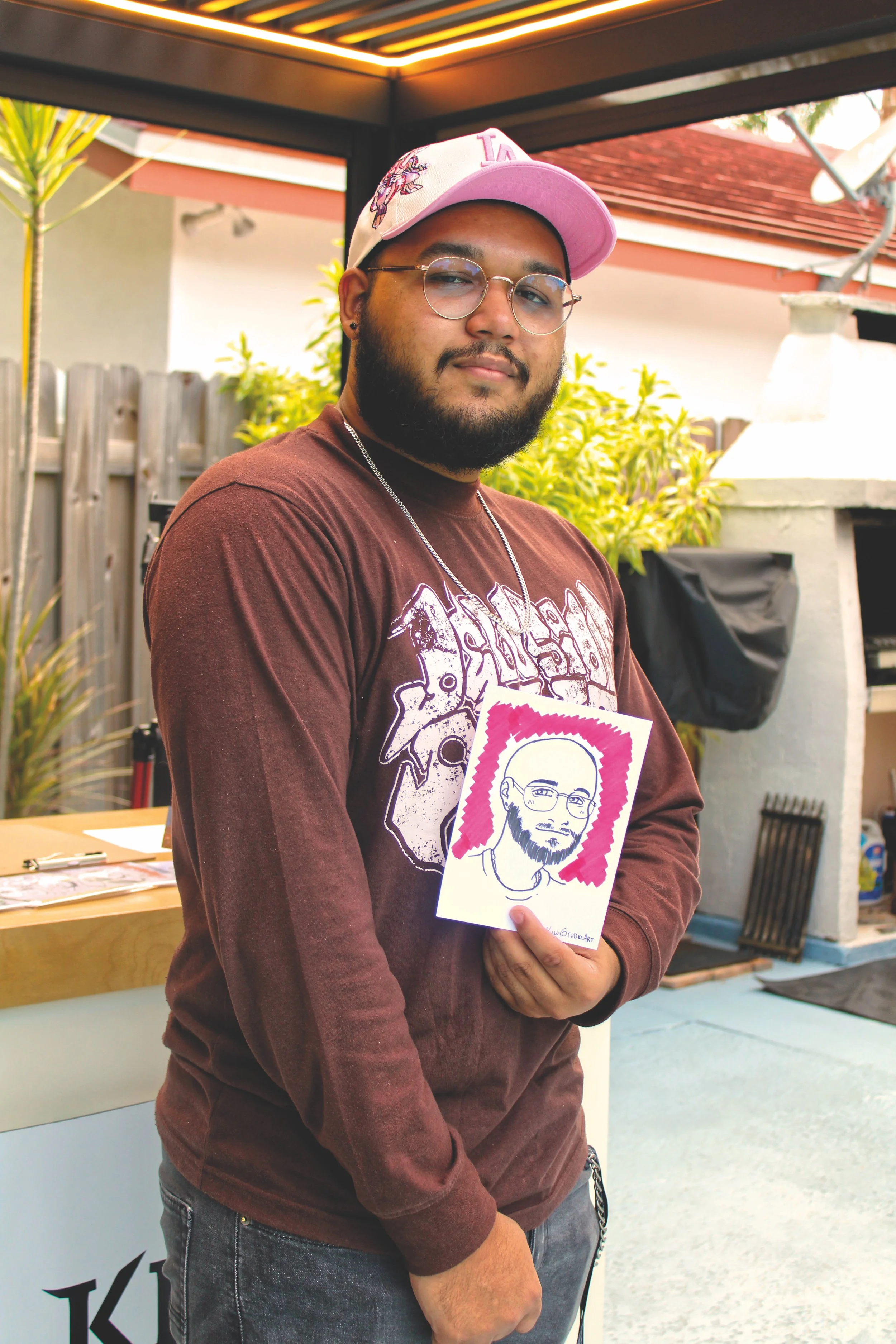 A bearded man wearing glasses and a pink baseball cap with a unicorn design, holding a caricature portrait of himself, standing outdoors in a backyard with a grill, wooden fence, and plants in the background.