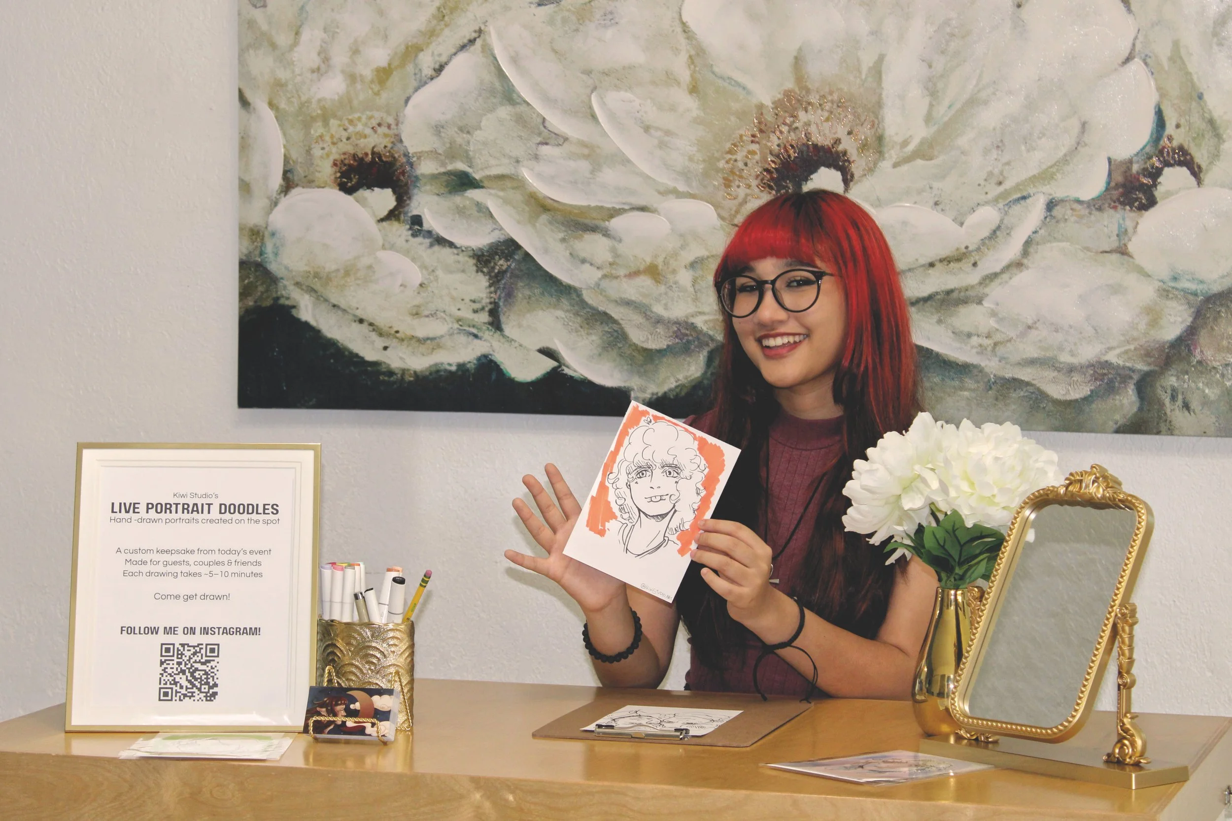 A woman with red hair and glasses smiling while holding a caricature portrait at an art event. She is standing behind a table displaying drawing supplies, a framed sign about live portrait drawings, and a QR code for social media. There is a large floral painting in the background.
