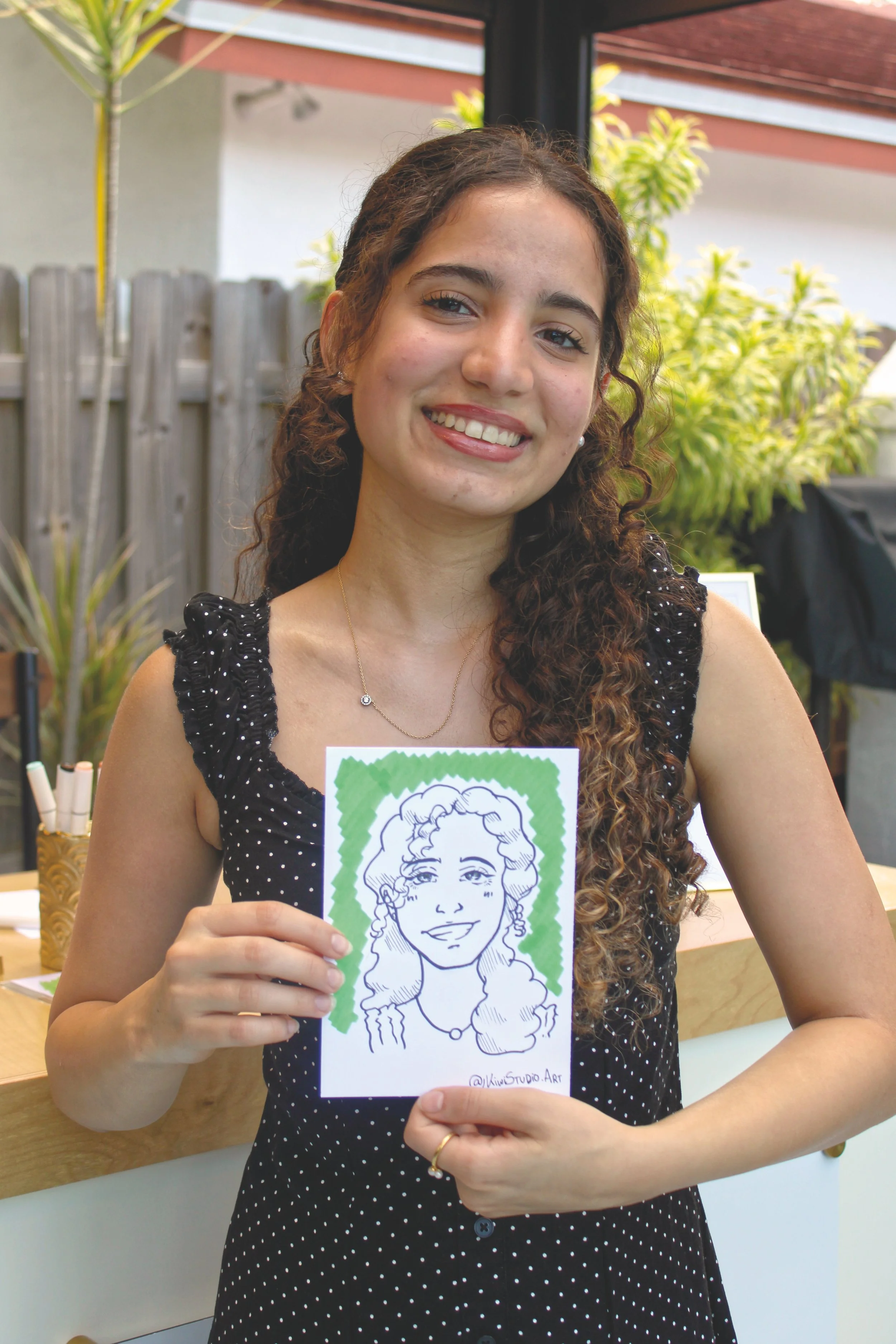 A young woman with curly brown hair and a black dotted dress holding an illustration of herself made by an artist.