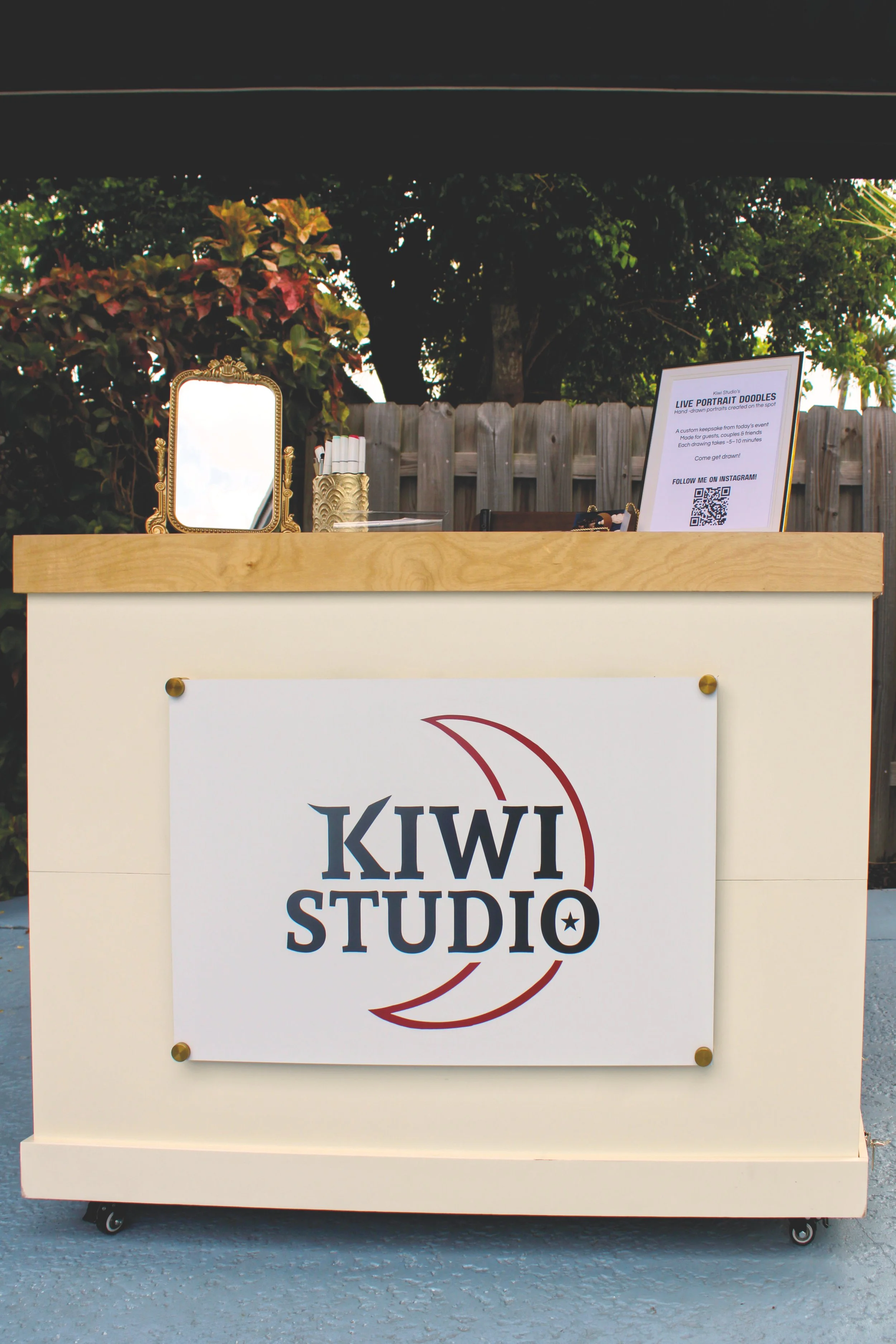 A white booth with a sign displaying "KIWI STUDIO" and a red crescent moon logo, set outdoors on a paved surface with a wooden fence and greenery in the background.