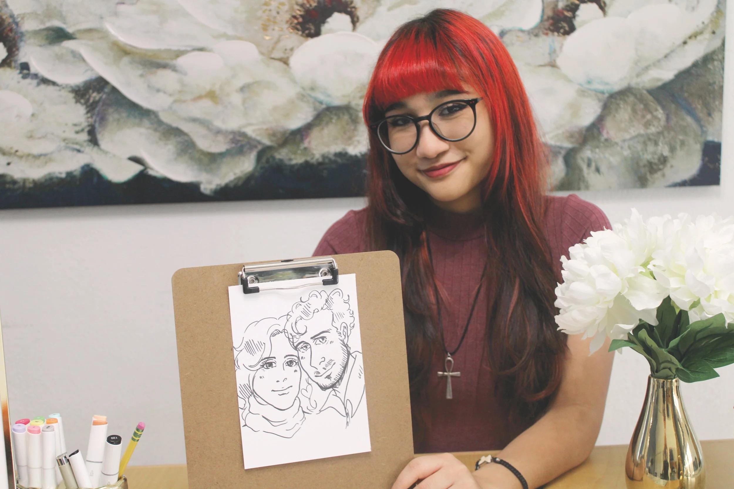 A young woman with red hair and glasses smiling at a desk with drawing supplies, holding an illustration of two people on a clipboard she made, and a vase of white flowers.