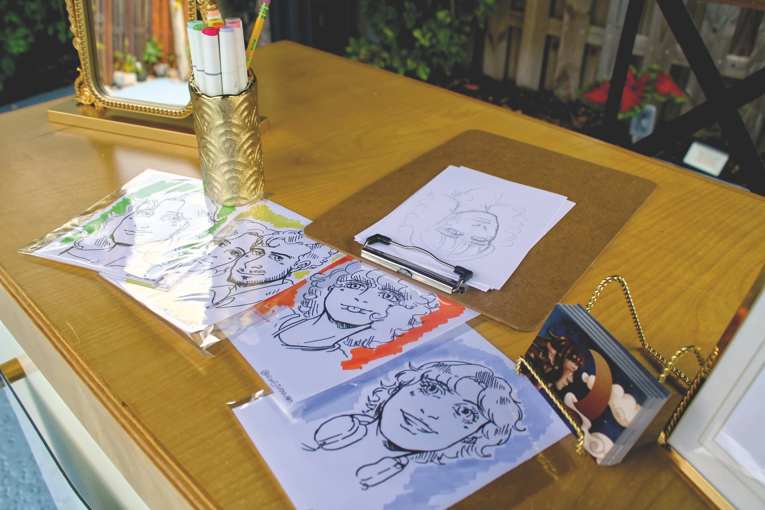 A wooden table holds several portraits of women and a man, some with colored backgrounds, along with a clipboard and sketch pages. A gold vase with colorful markers, a mirror with a decorative gold frame are also visible on the table. The background shows an outdoor setting with a wooden fence and some greenery.