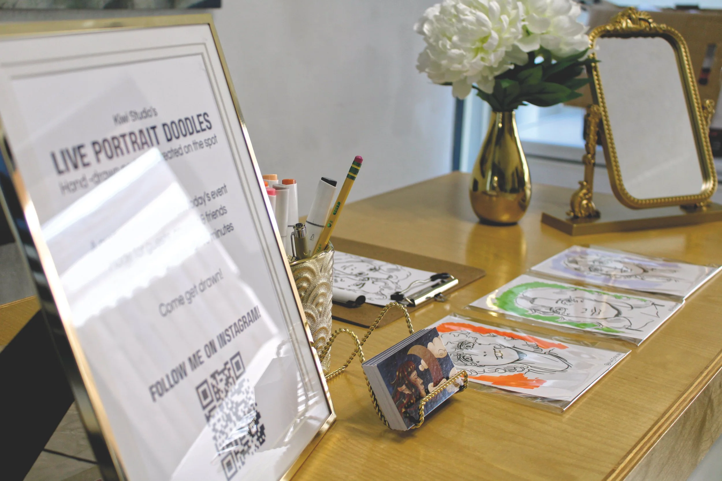 A table displays artistic prints and drawings, a gold flower vase with white flowers, a gold-framed mirror, and a sign with a QR code, advertising live portrait doodles.