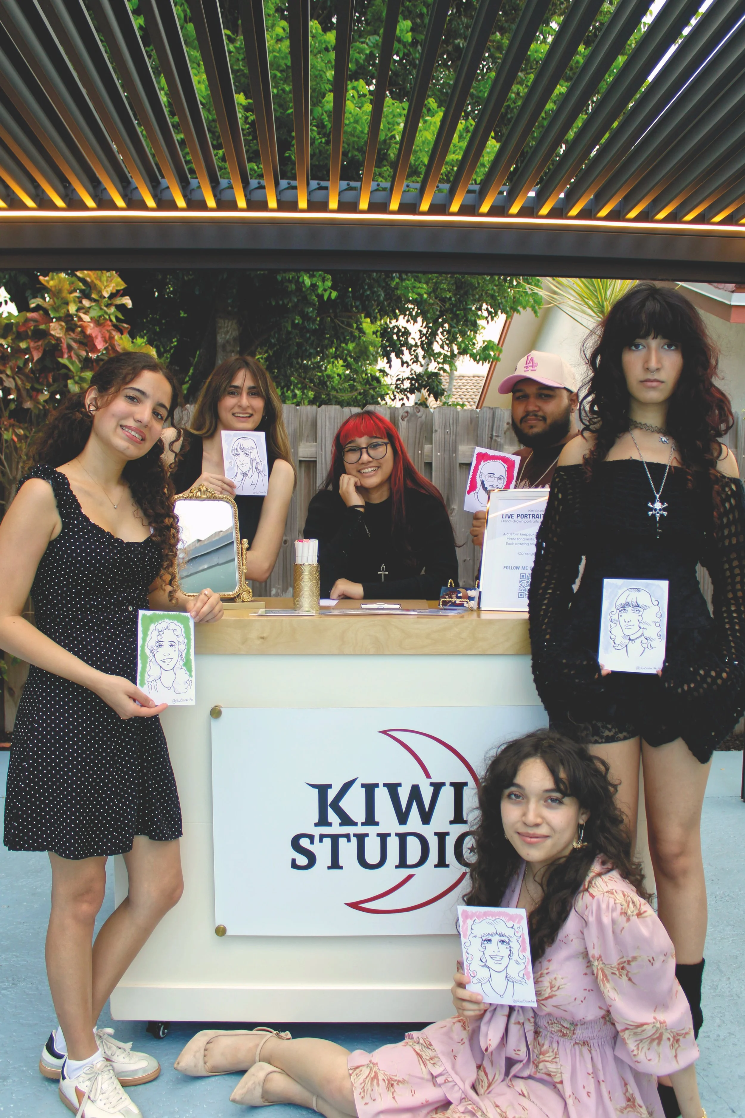 Group of happy guests at a booth, holding illustrated portraits of themselves, with a sign that reads 'KIWI STUDIO' and a fence and trees in the background. The artist in the middle smiling while the others show the portraits she made them