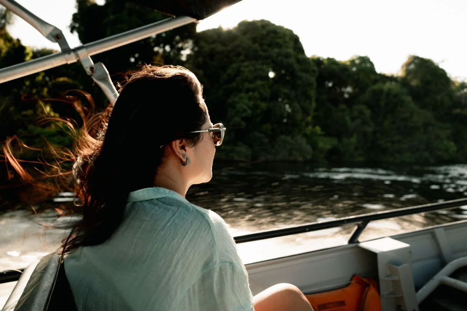 Mulher com cabelos ao vento, usando óculos de sol, sentada em um barco em um rio com árvores ao fundo, ao pôr do sol.