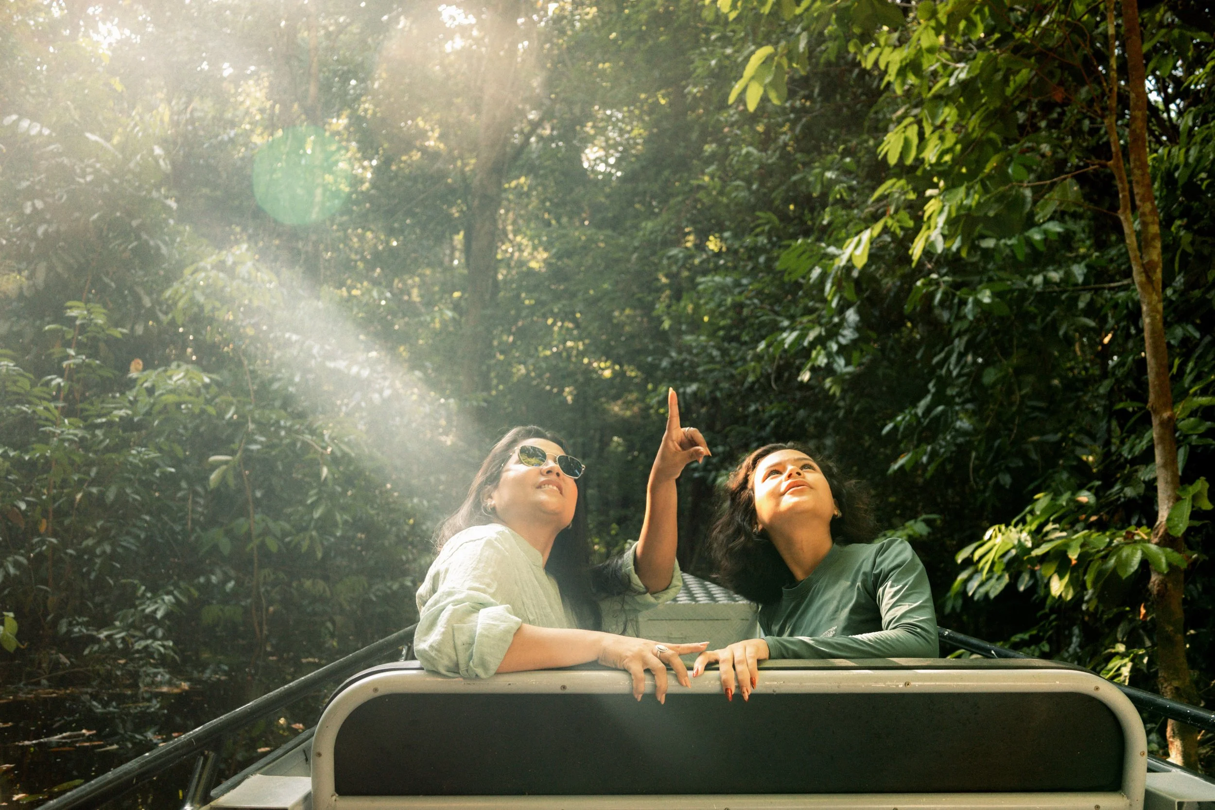 Duas mulheres sentadas na parte de trás de um veículo em uma floresta tropical, uma delas apontando para as árvores acima, com luz do sol filtrando através das folhas.
