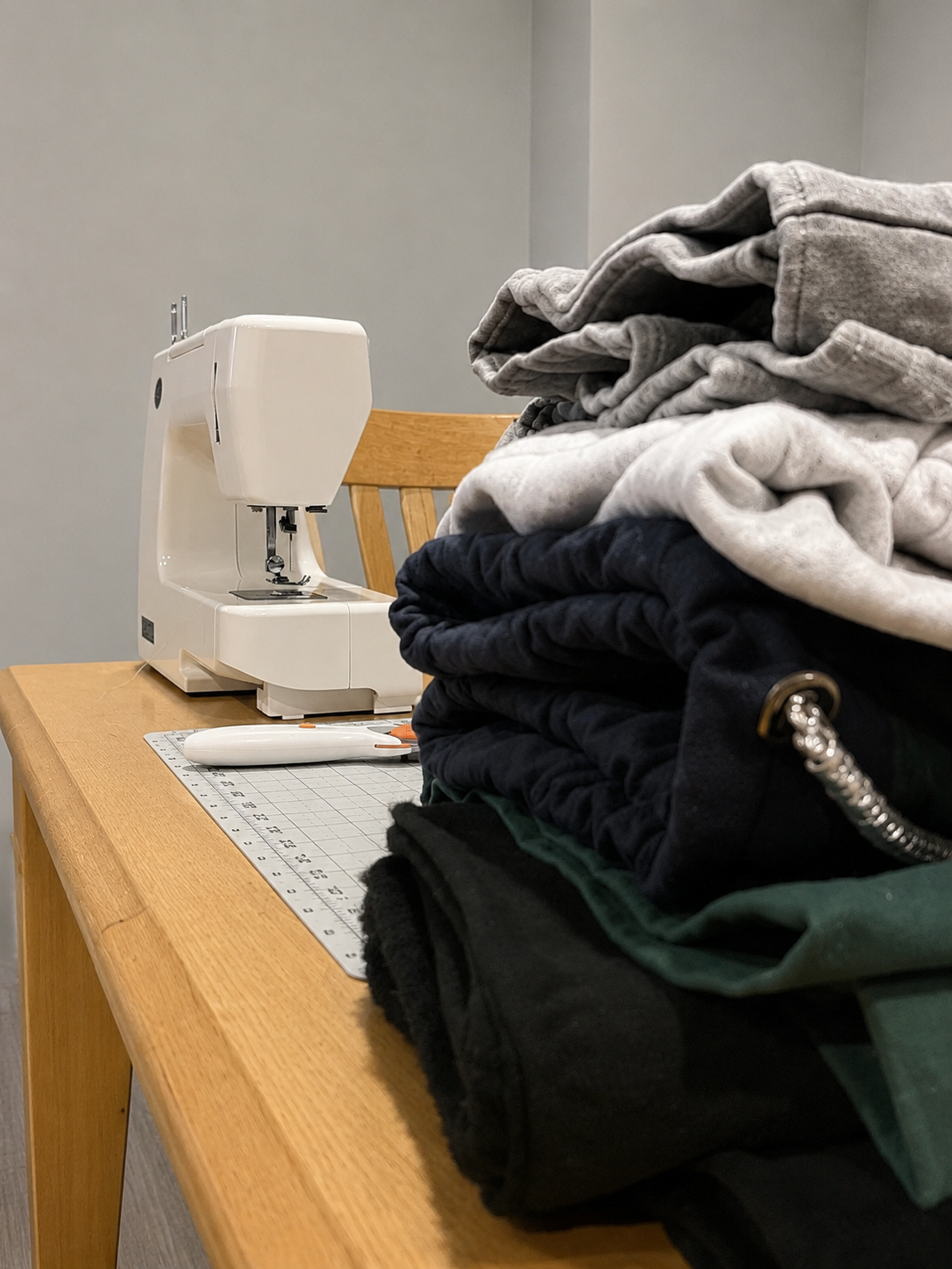 A pile of folded gray, black, and green clothing on a wooden table, with a white sewing machine and a cutting mat with a rotary cutter nearby.