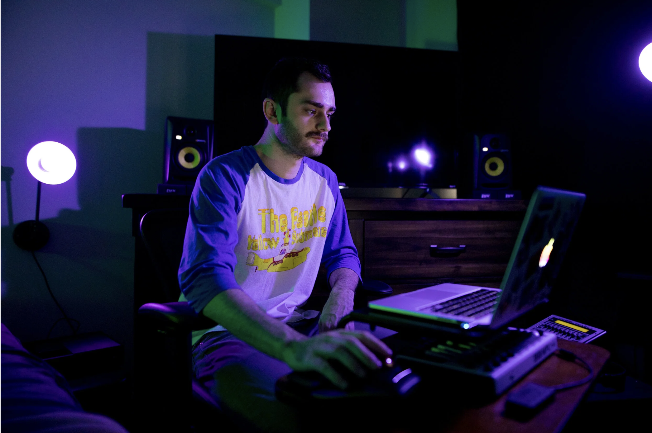 A young man with dark hair and a mustache sits at a desk using a laptop, surrounded by purple and green ambient lighting in a dark room. He is wearing a white and blue raglan shirt with yellow text and graphics.