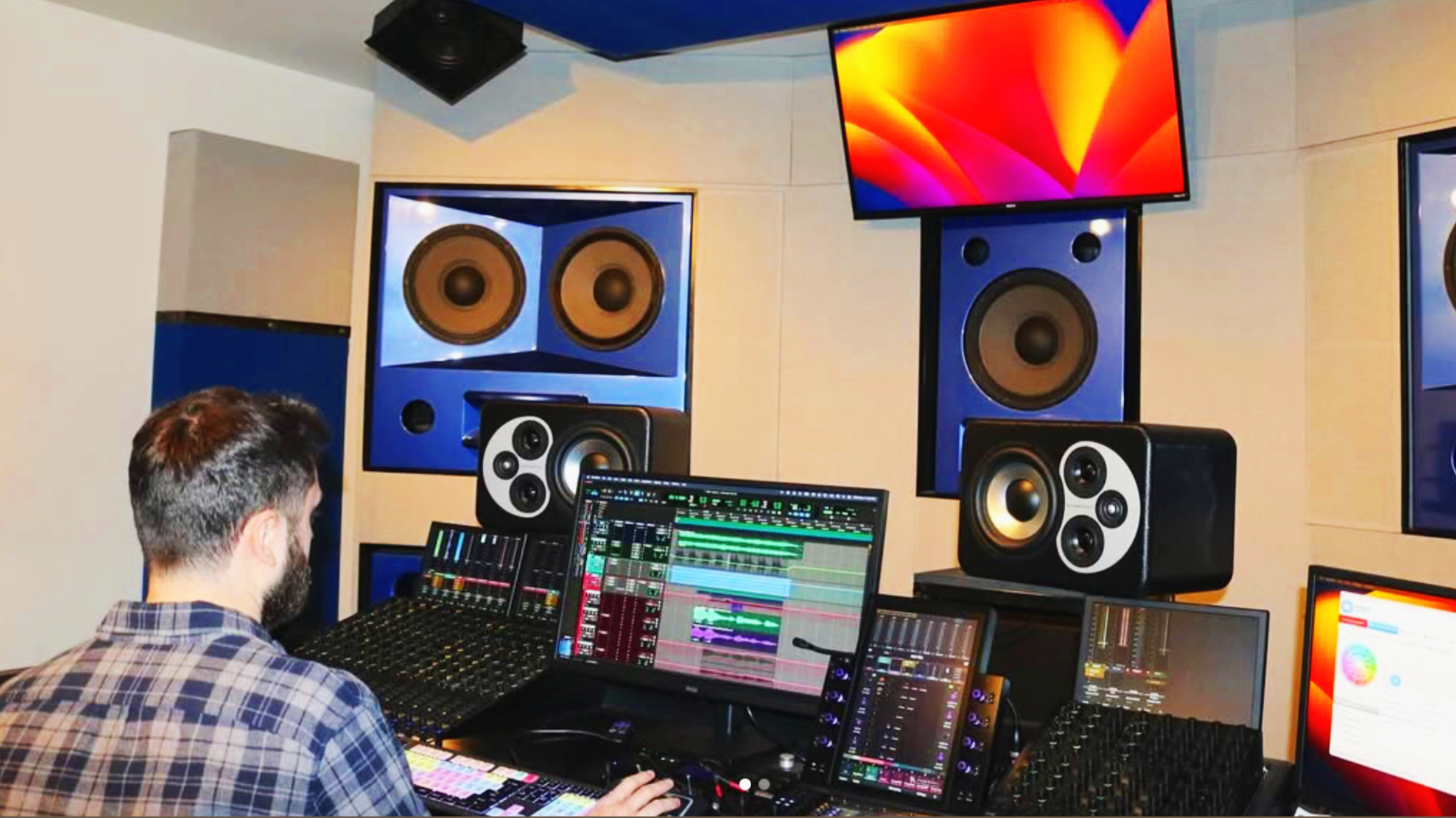 A person working in a music studio with multiple computer monitors displaying audio editing software and four large speakers mounted on the wall.