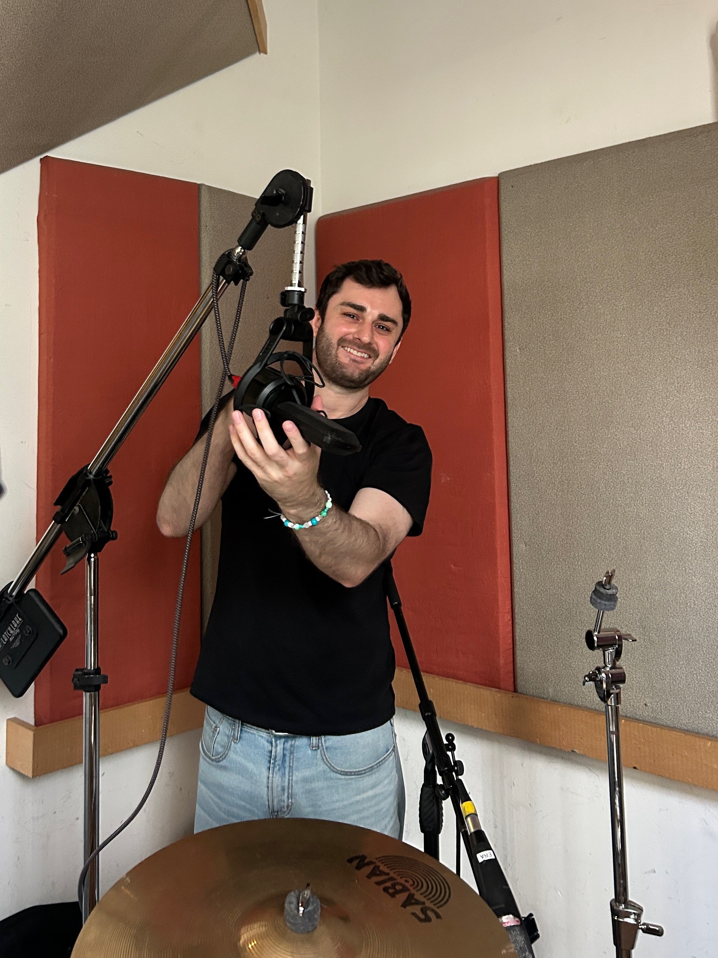 A man smiling in a recording studio, holding headphones, surrounded by audio equipment and soundproof wall panels.