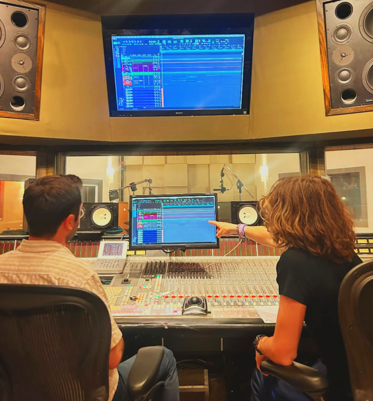 Two people in a recording studio control room working with audio mixing equipment and computer screens displaying digital audio workstations. One person is pointing at the screen while the other listens.