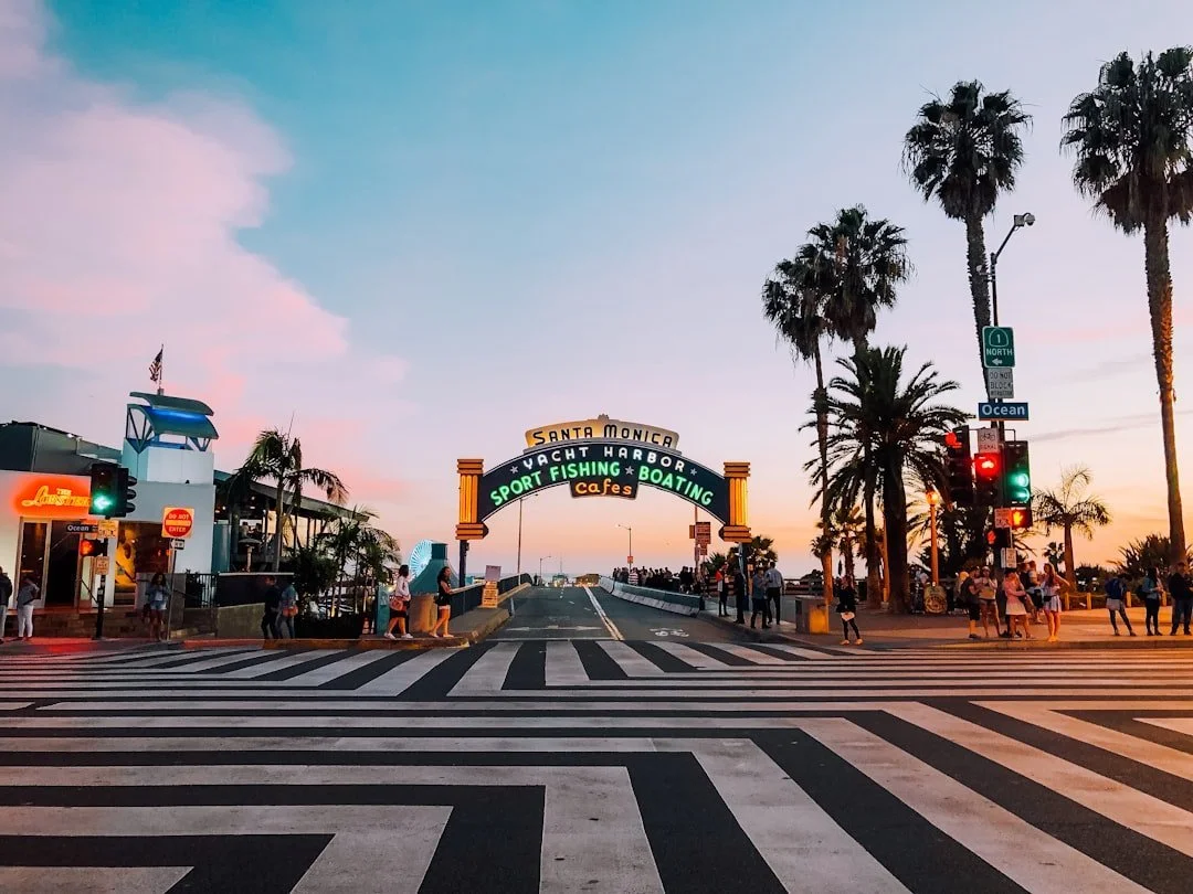 Street view at Santa Monica Pier in California during sunset, with palm trees, a sign for sport fishing, boating, cafes, and the Pacific Ocean in the background.